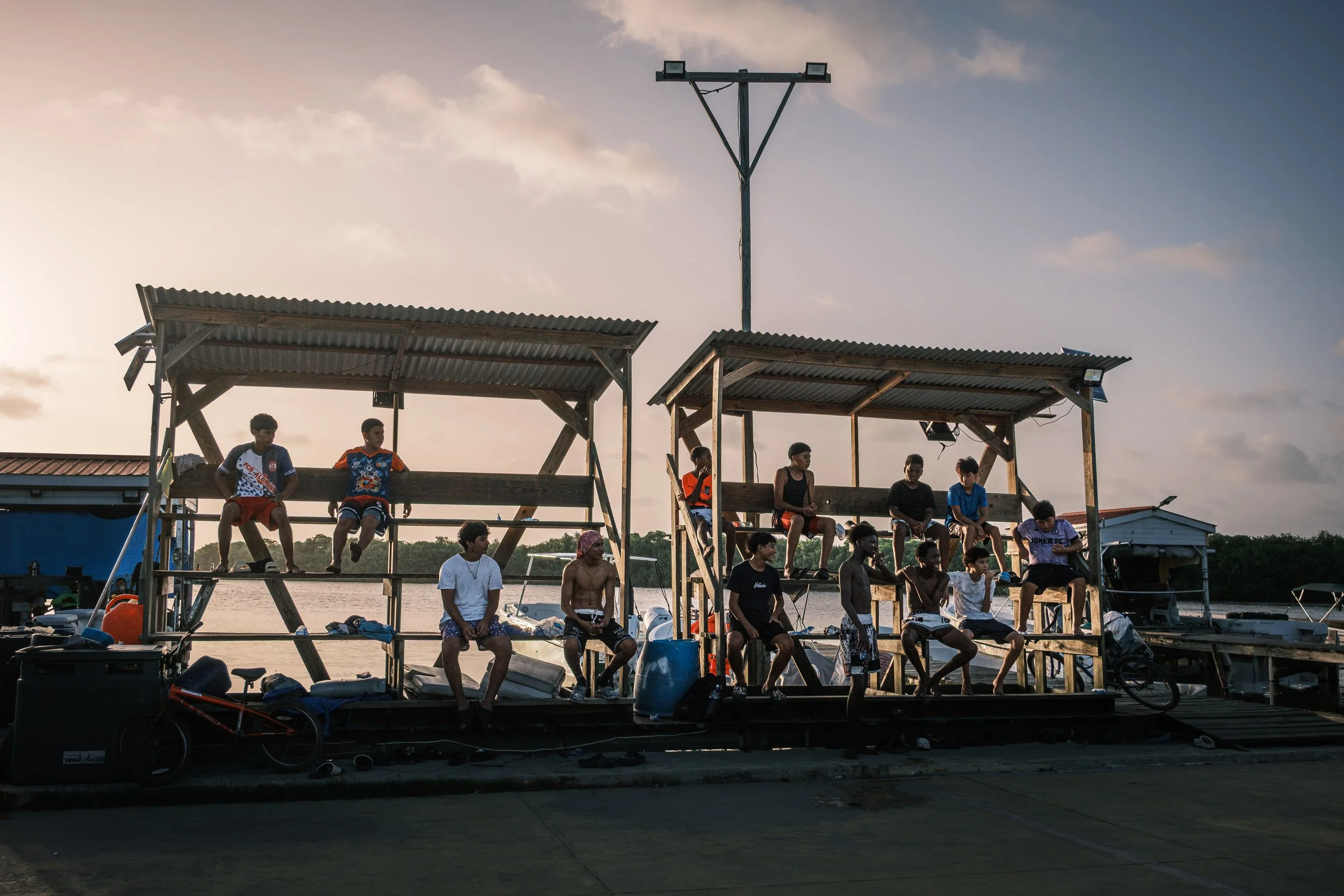 A group of teenagers sitting on a dock by the water during sunset, some wearing life jackets and casual clothing, with various items and boats around them.