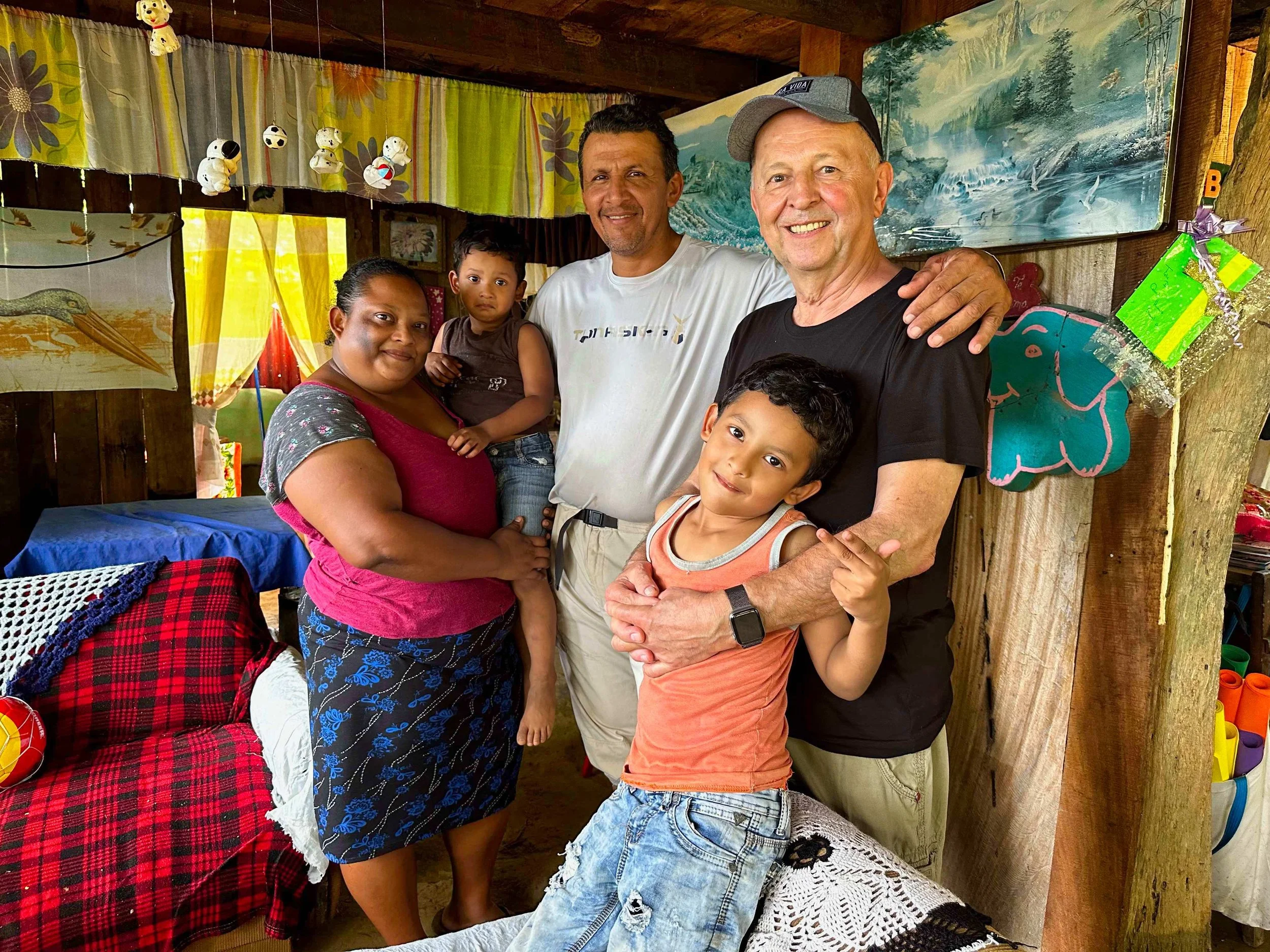 A group of five people, two adults and three children, posing together inside a rustic room with wooden walls, colorful curtains, and decorations. They are smiling and appear happy.