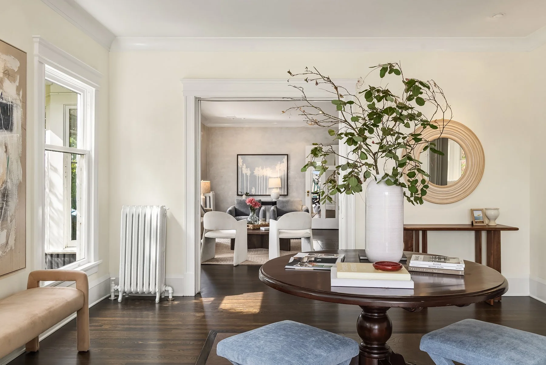 Staged dining area with a round wood table and fresh branches, opening into a bright living room