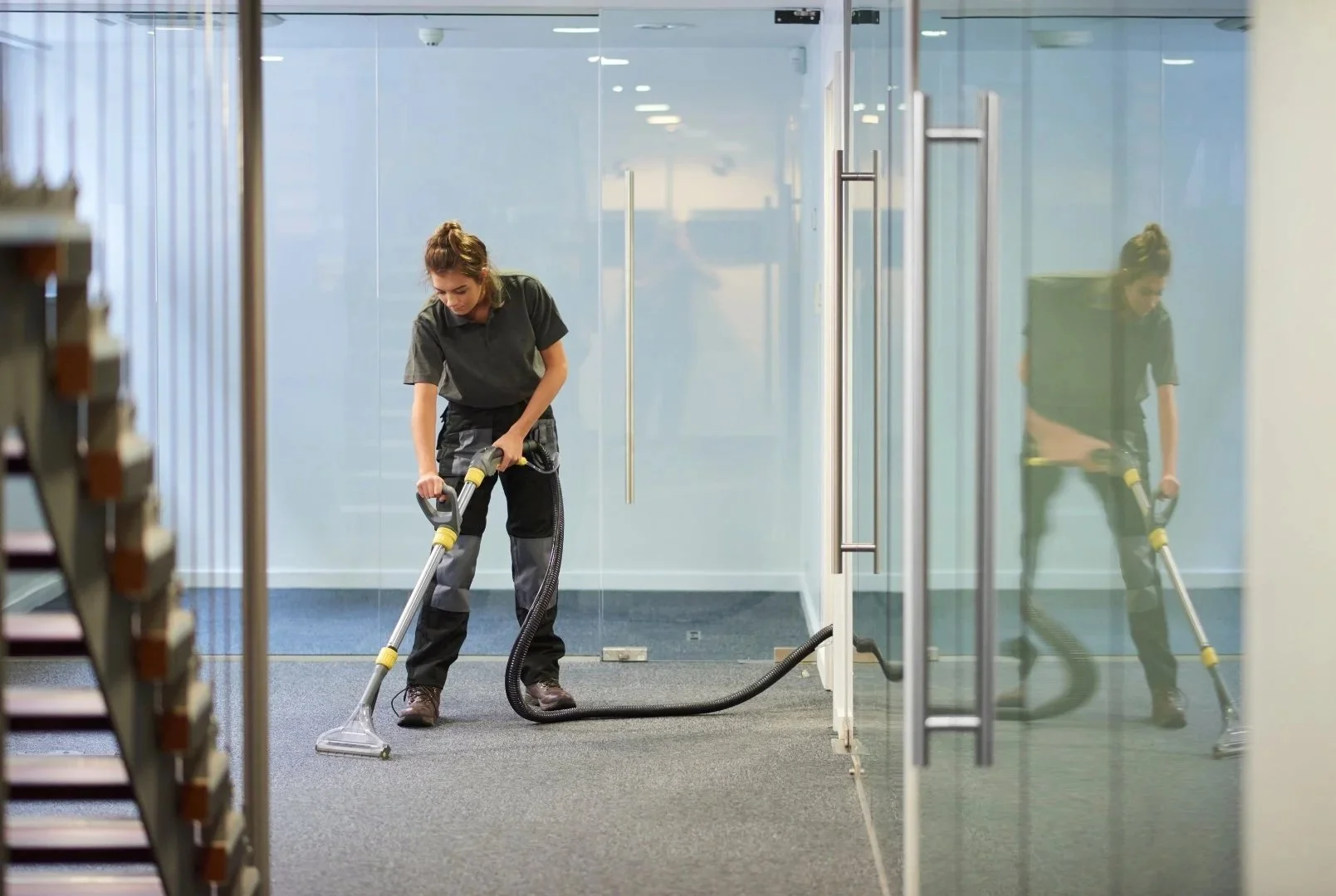 A woman cleaning a carpet with a commercial vacuum cleaner in an office hallway.