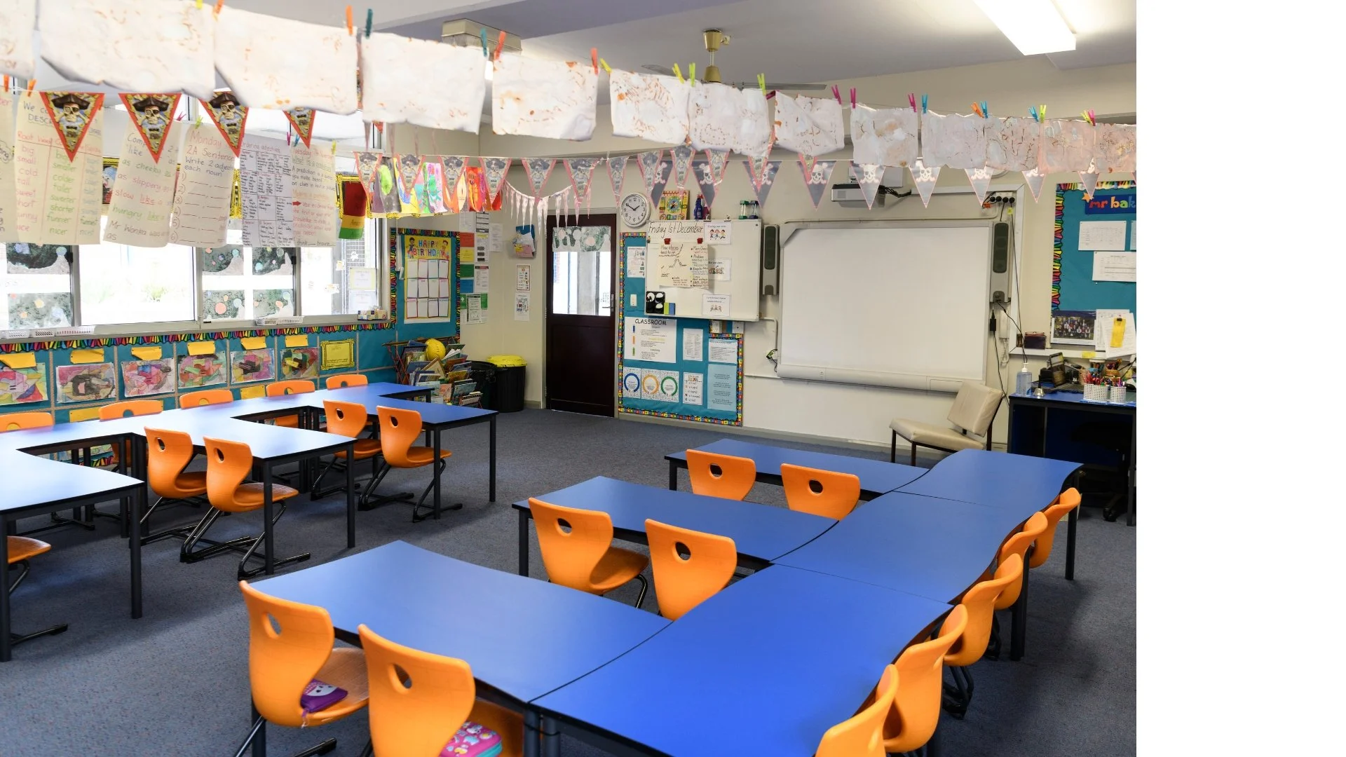 An empty classroom with orange chairs, black tables, a whiteboard, colorful posters, and decorations hanging from the ceiling.