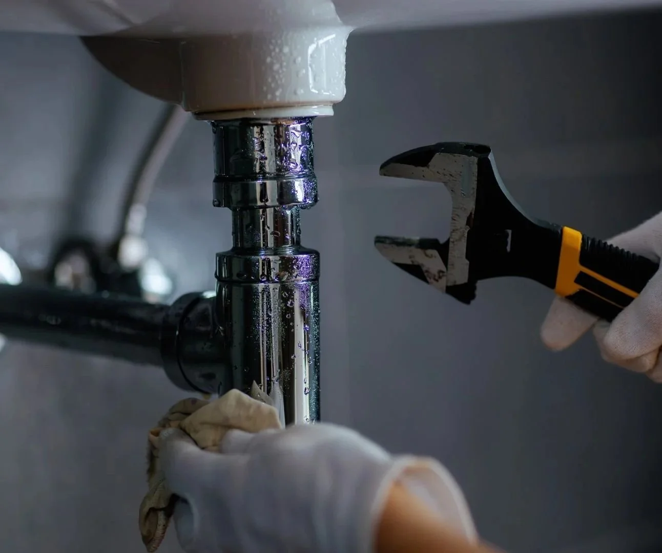 A person wearing white gloves using a wrench to tighten a pipe under a sink, with water droplets on the pipe.