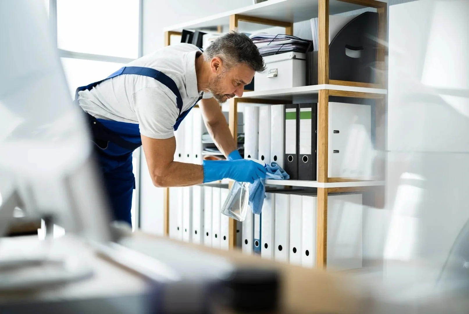 A man wearing a white shirt, blue apron, and blue gloves cleaning a shelf with black and white binders in an office.