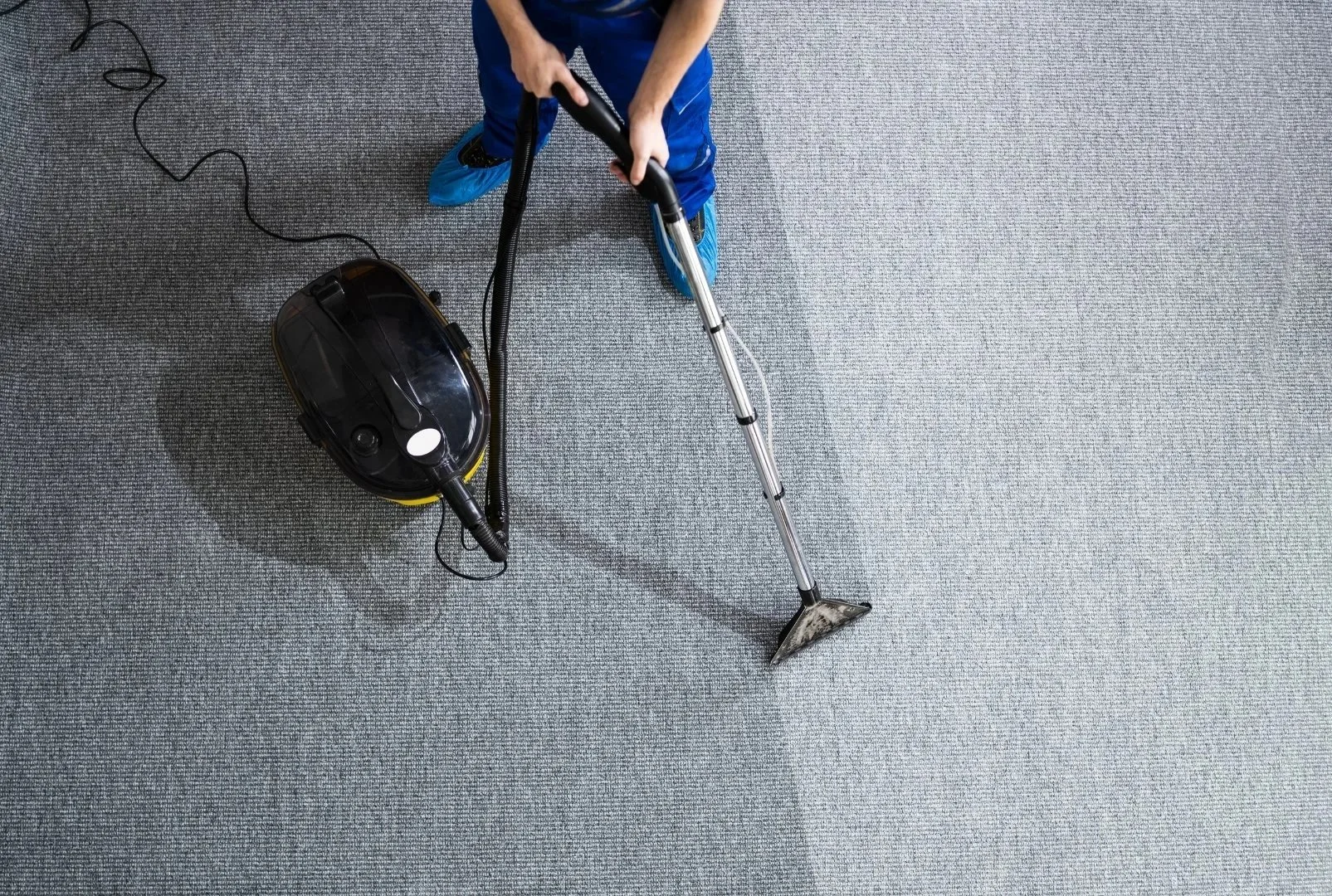 Person using a vacuum cleaner on a gray carpeted floor, wearing blue shoes and blue clothing.