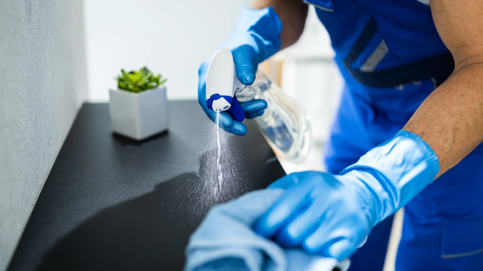 Person wearing blue uniform and gloves disinfects a surface with spray disinfectant.