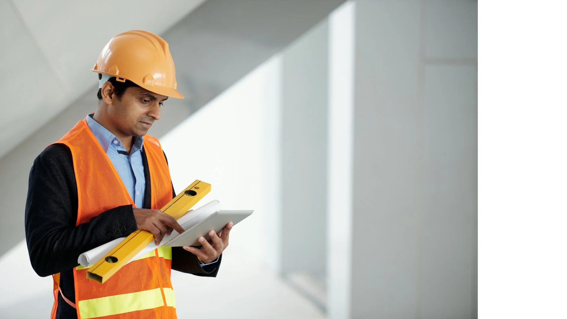 Construction worker in safety vest and helmet using a tablet and level in an indoor site.