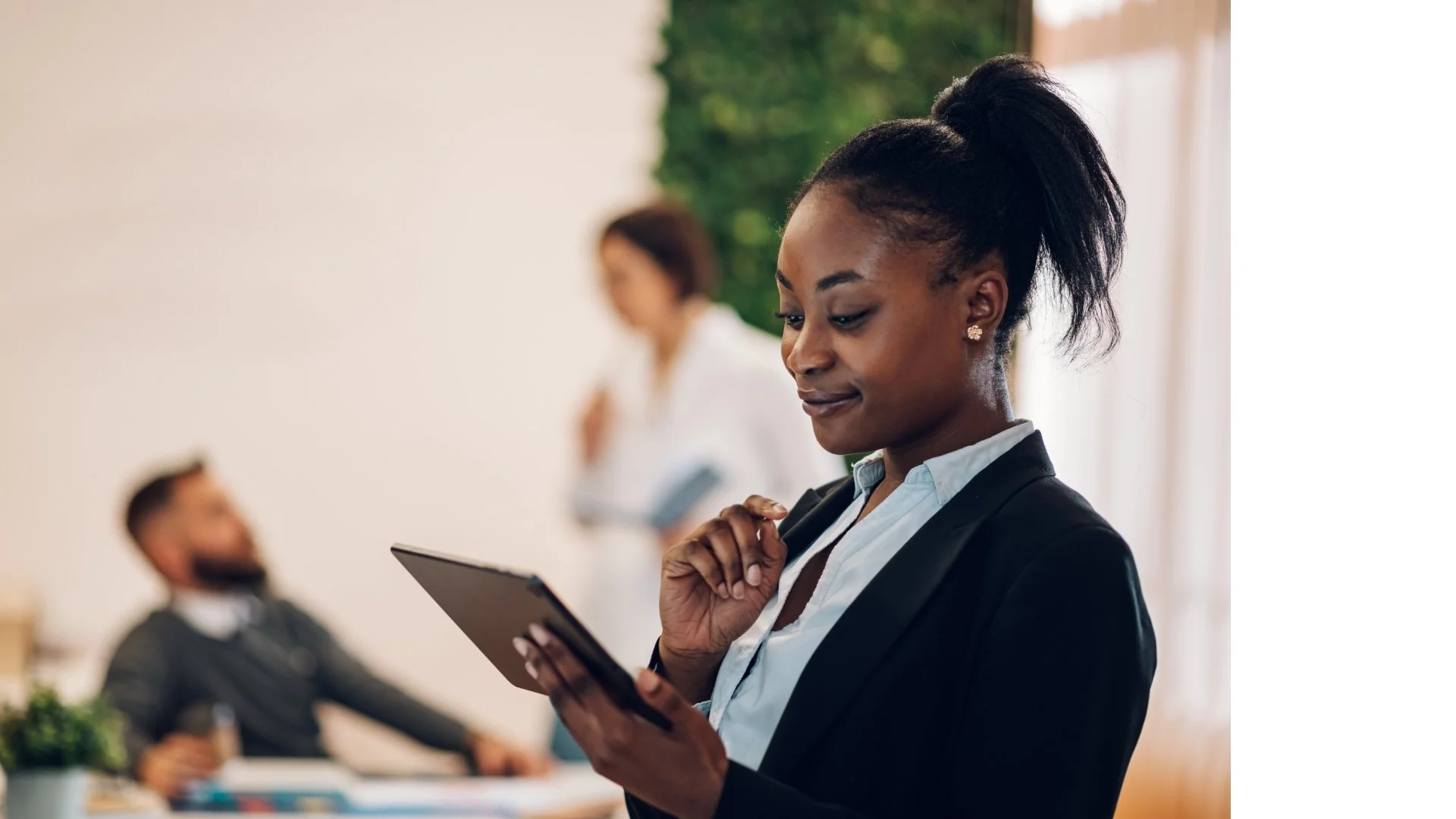 Businesswoman in a black blazer looking at her tablet in an office, with a man and woman working in the background.