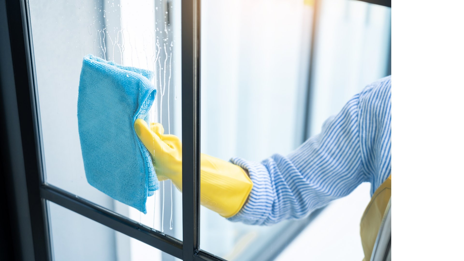 Person cleaning glass window with a blue cloth, wearing yellow rubber gloves and a striped long-sleeve shirt or uniform.