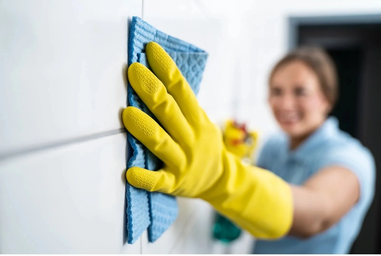 Person in a blue shirt cleaning white tiled wall with a yellow glove and blue cloth, smiling.