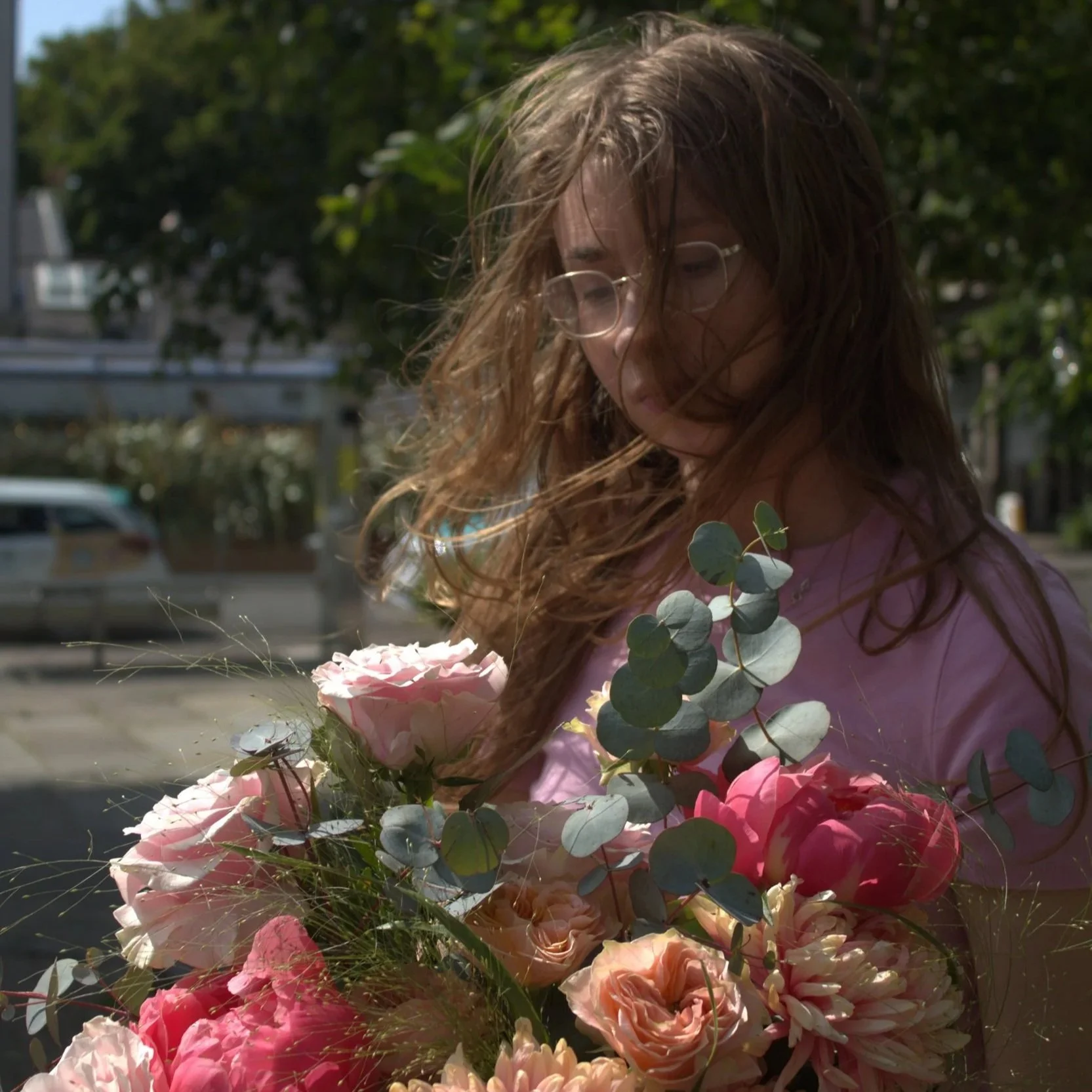 A woman with long, wavy brown hair and glasses holding a large bouquet of pink and peach flowers with eucalyptus leaves outdoors on a sunny day.