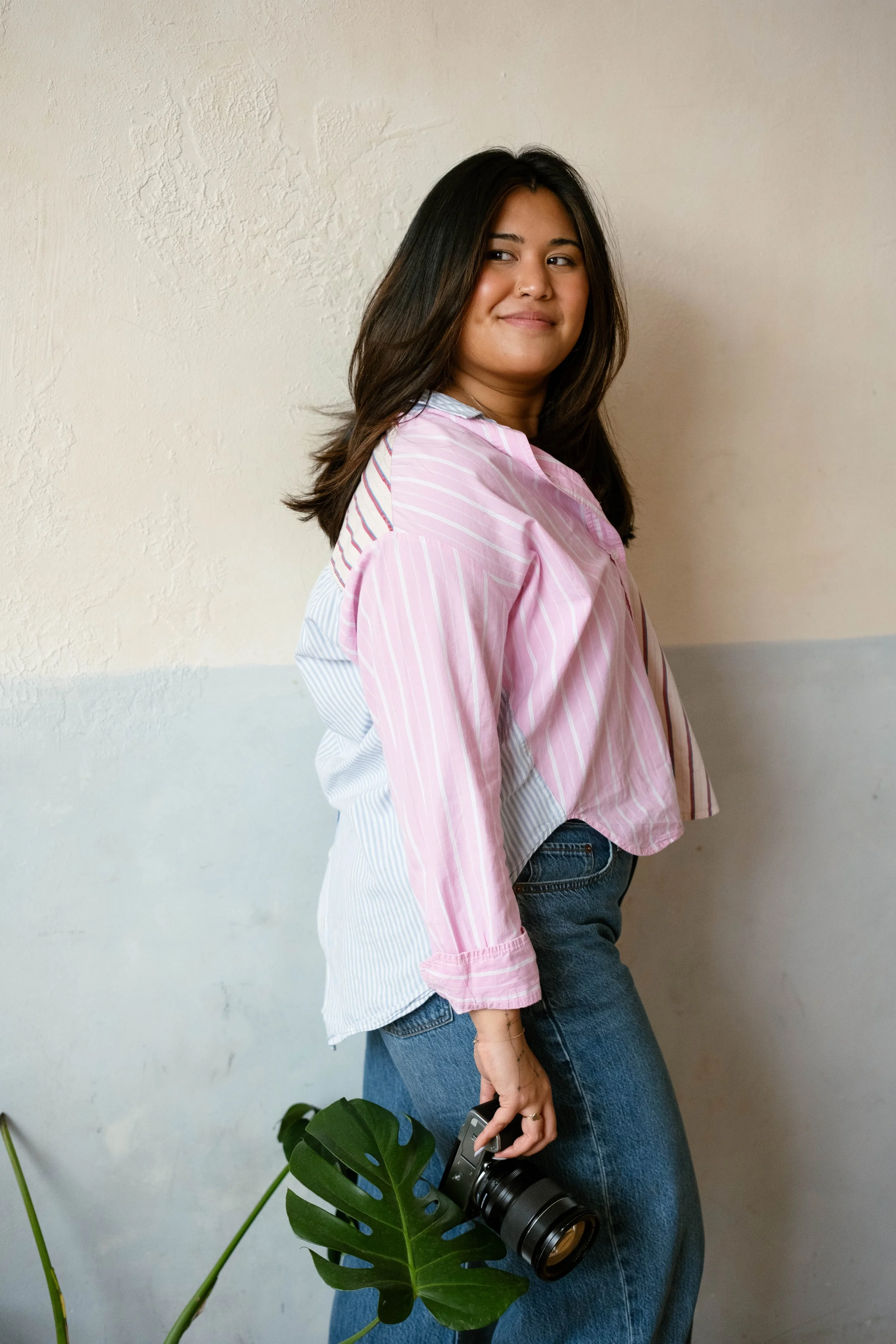 A young woman with dark hair, wearing a pink and white striped shirt and blue jeans, standing indoors next to a white wall, holding a black camera with a large lens, with a green plant in the foreground.