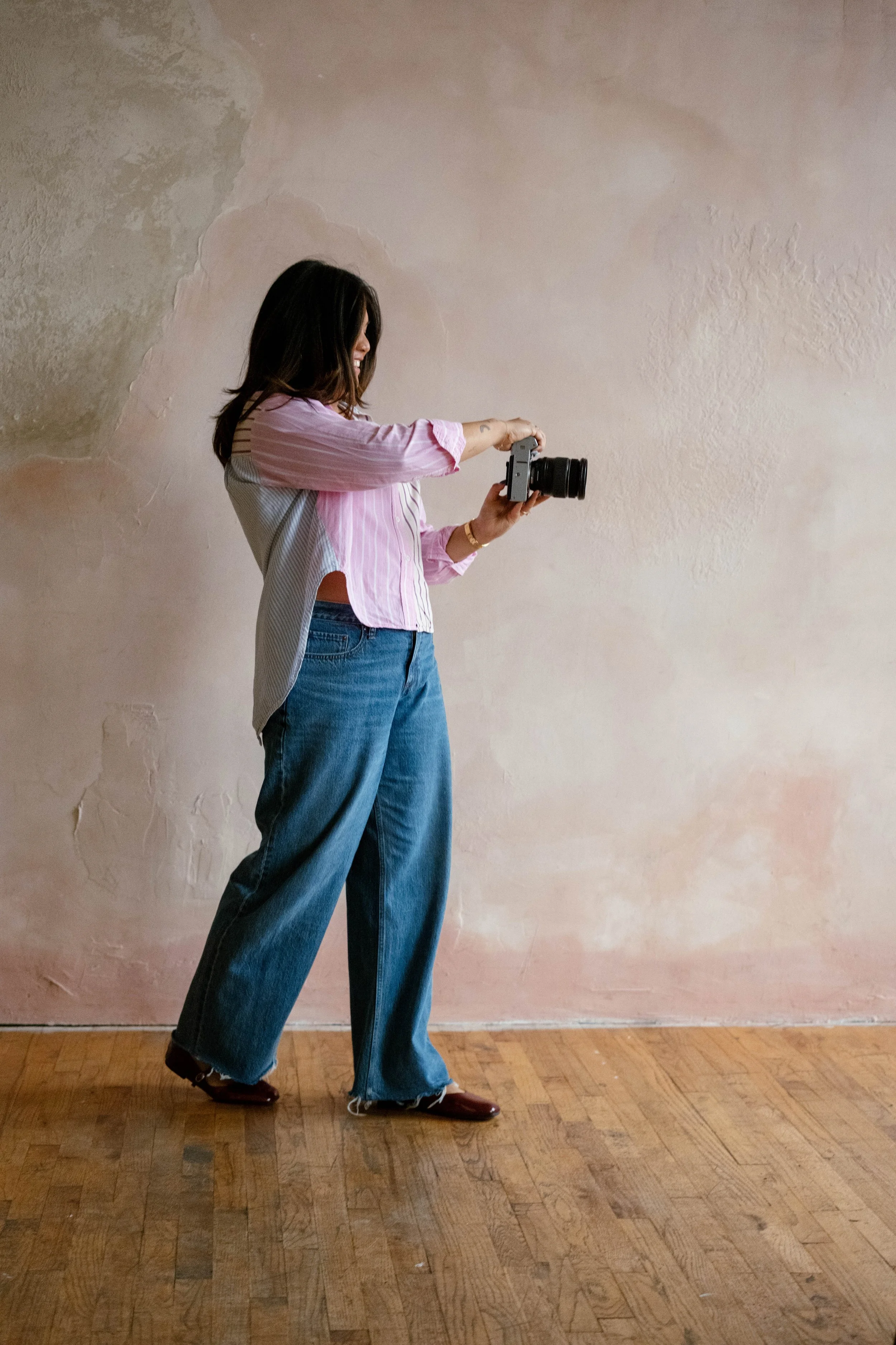 A young woman with dark hair, wearing a pink and white striped shirt and blue jeans, is standing on a wooden floor, holding a camera while taking a picture. The background is a textured, partially faded pink wall.