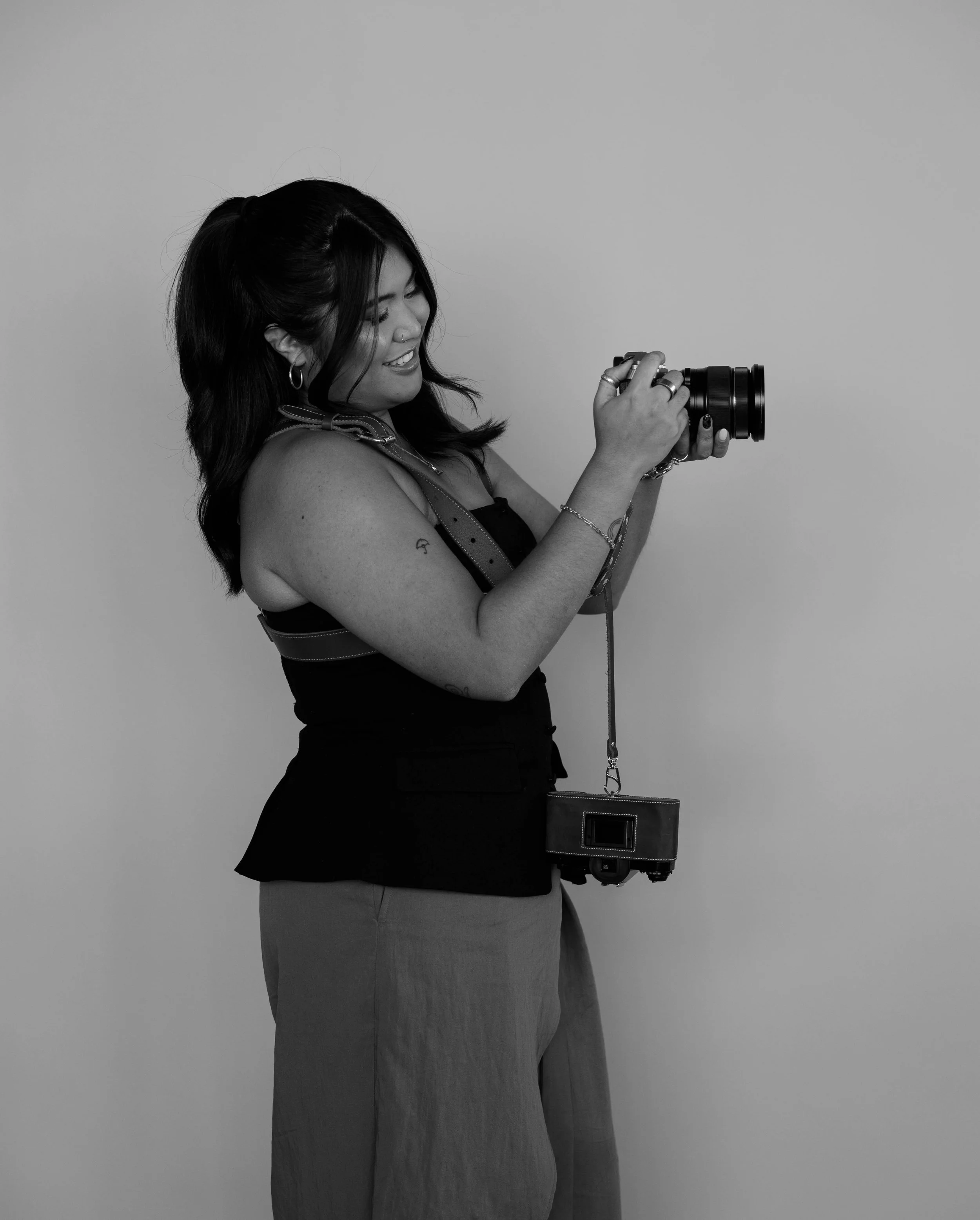 A woman with medium-length dark hair and tattoos, smiling and holding a camera, standing against a plain background.
