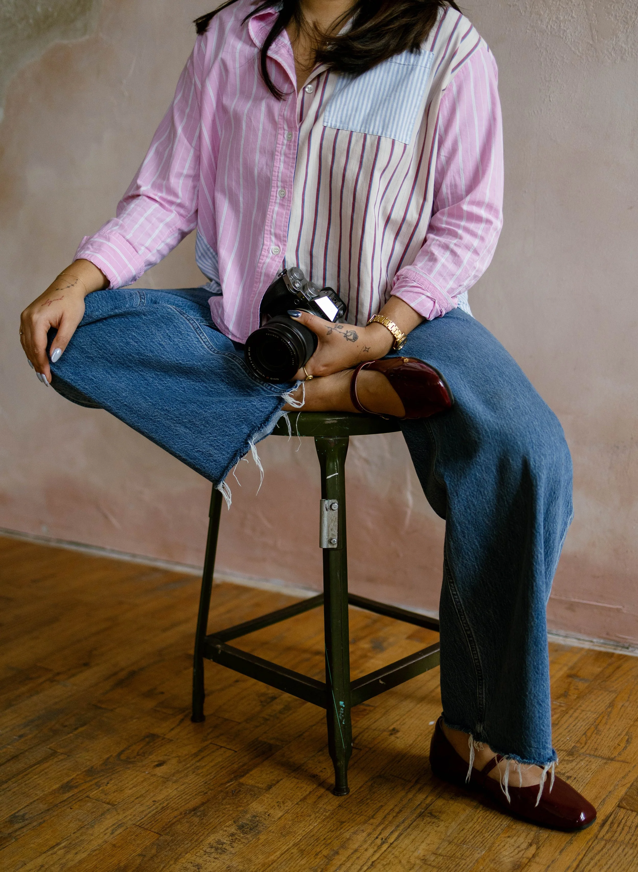 Person sitting on a green metal stool with legs crossed, holding a camera, wearing a pink and blue striped shirt, frayed jeans, and brown flats, against a beige wall and wooden floor.