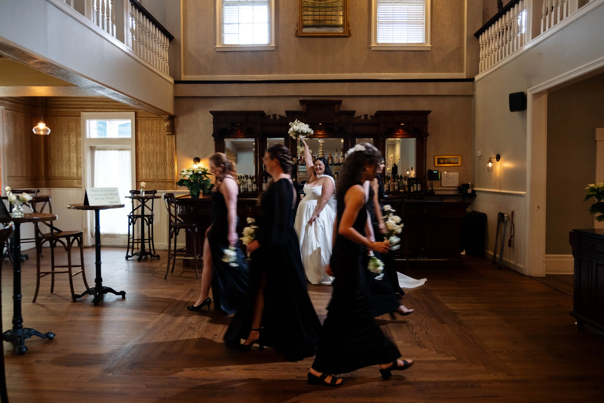 Bridal party dancing in a wedding reception hall, with a bride in a white dress holding a bouquet and smiling, surrounded by bridesmaids in black dresses holding flowers.