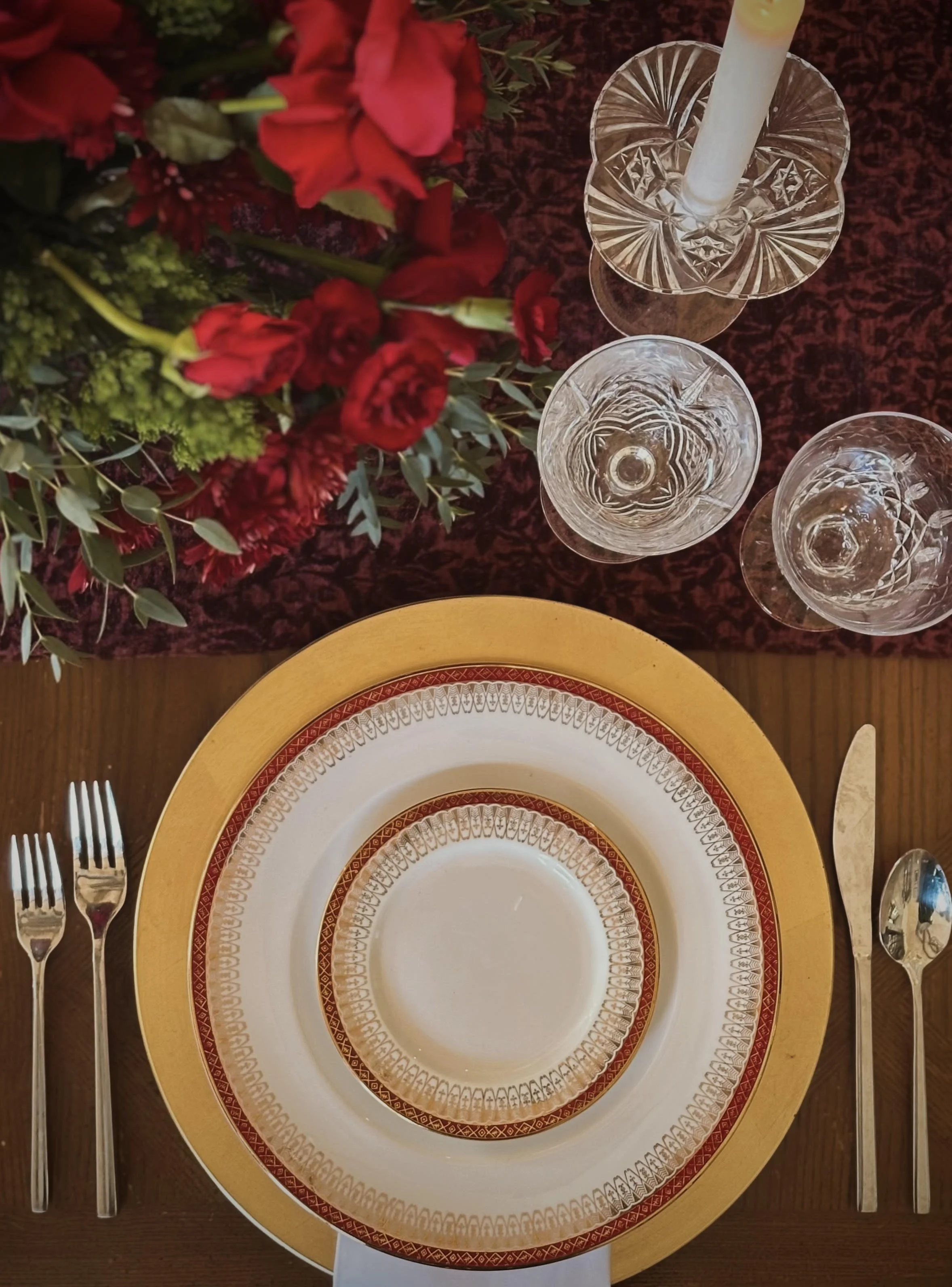 Elegant table setting with a large yellow charger plate, smaller patterned dinner plate, and a smaller patterned salad plate, accompanied by two forks on the left and a knife and spoon on the right. A flower arrangement with red and green flowers and foliage is in the top left corner, and three cut crystal glasses with intricate designs are positioned at the top right on a dark red cloth.