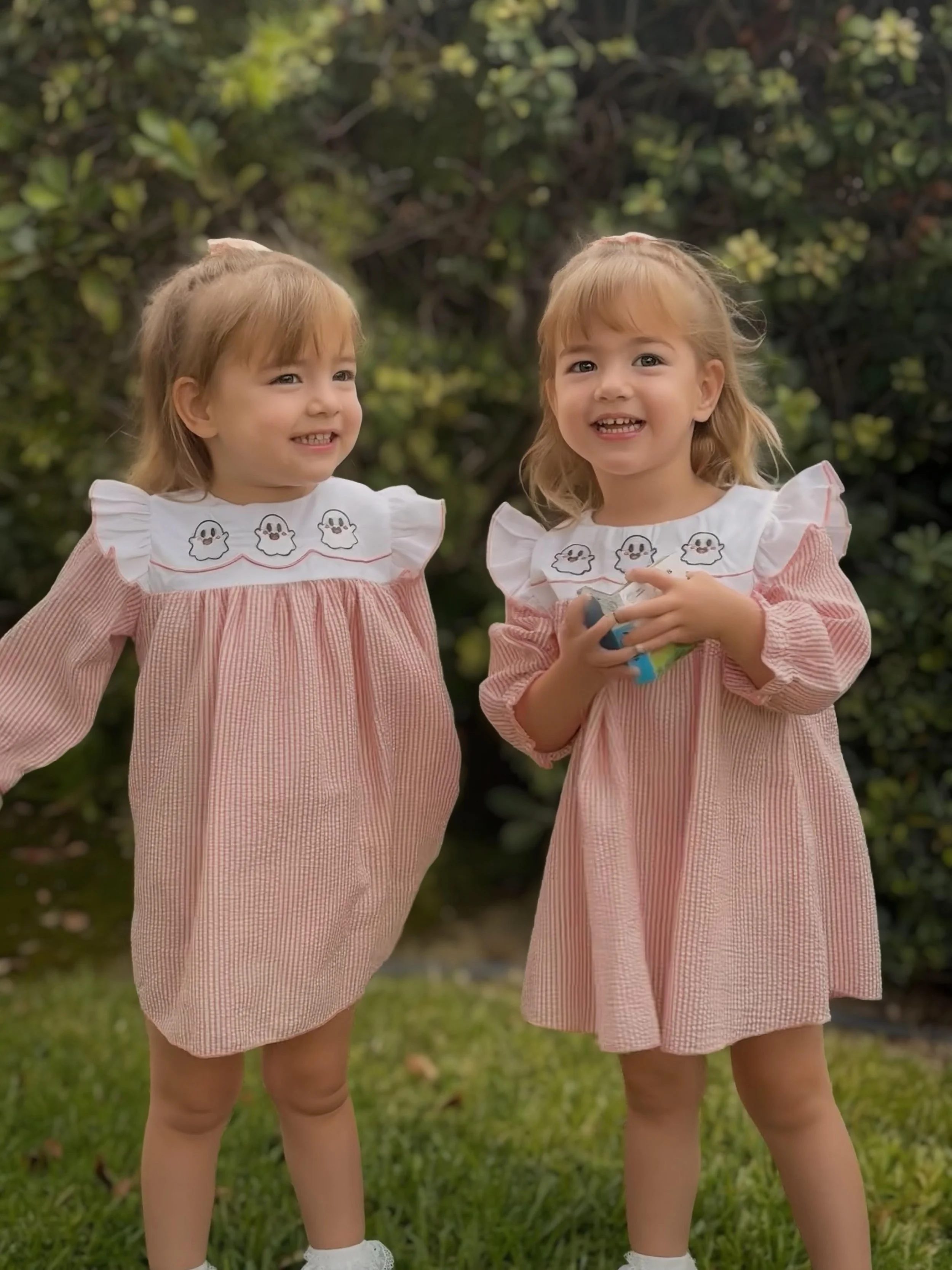 Two young girls with blonde hair, wearing matching pink and white dresses with ghost designs on the collar, are standing outdoors in front of green bushes. They are smiling and appear happy.