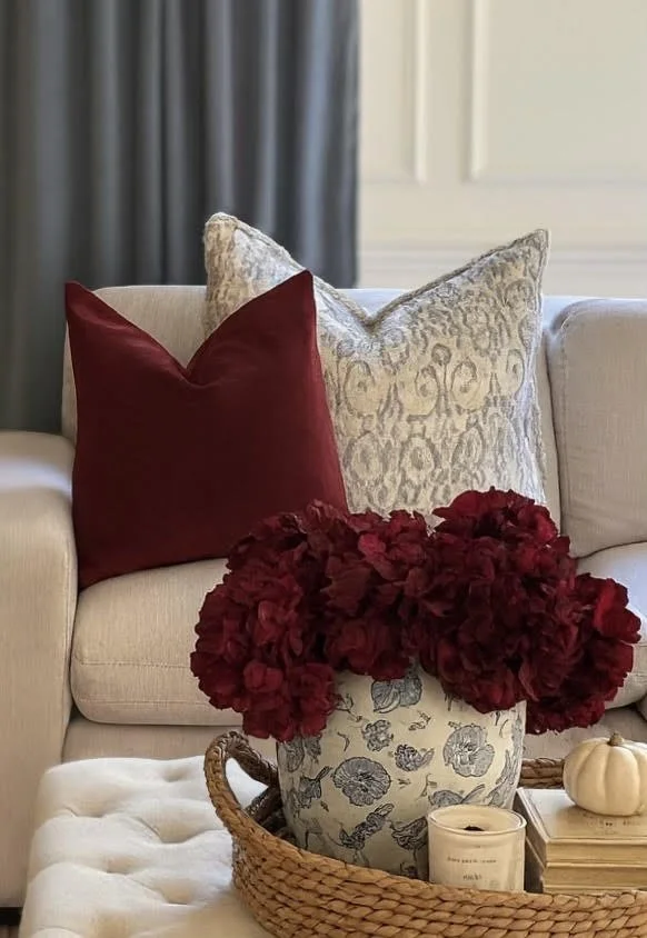 A living room scene with a beige sofa decorated with two pillows, one in burgundy and one in cream with decorative patterns. In the foreground, there is a vase with red hydrangeas, placed on a woven tray along with a lit candle, a small pumpkin, and a few books.