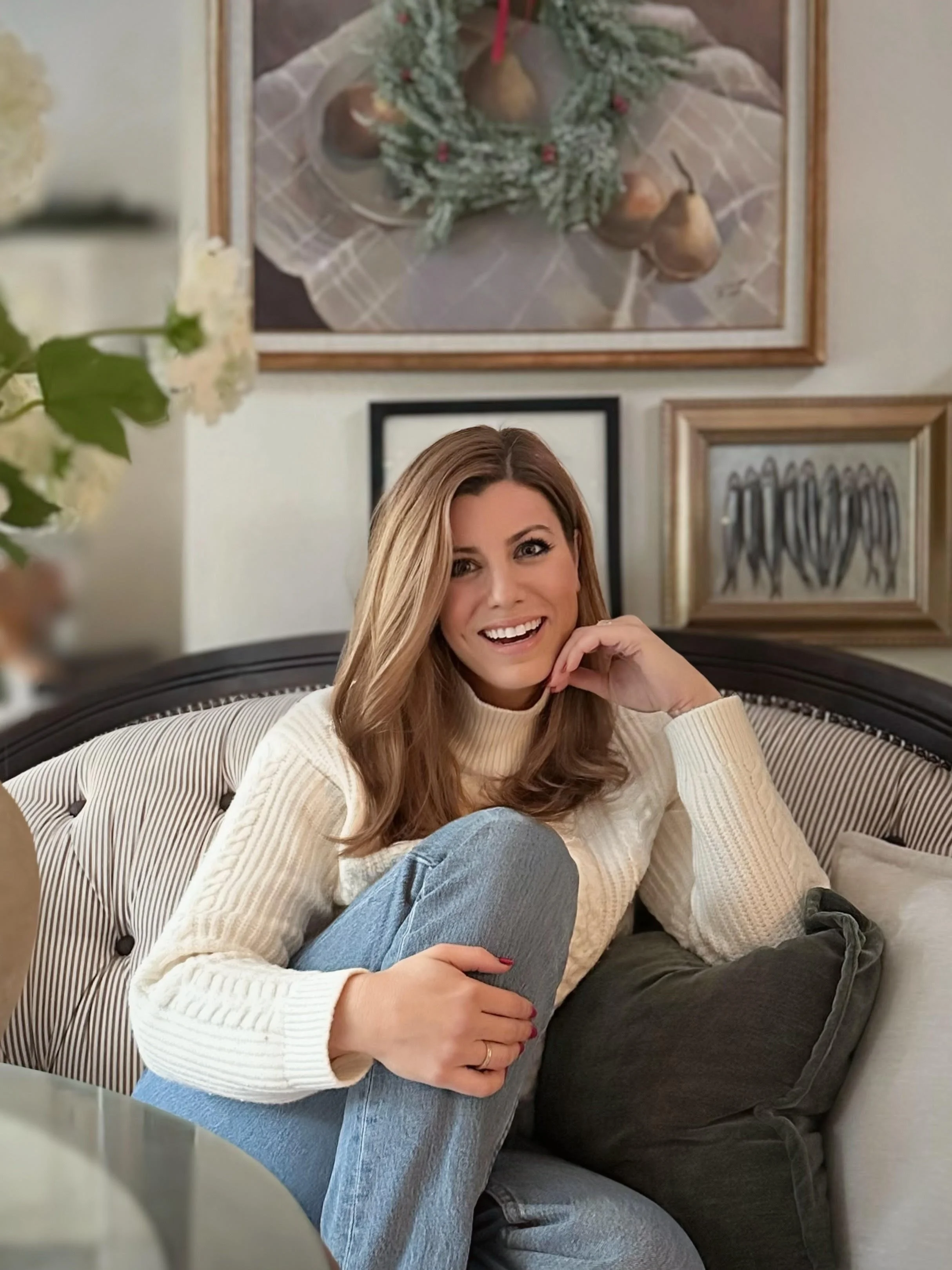 A woman with brown hair, smiling, sitting on a striped sofa with her knees up, in a cozy room. The background features framed artwork and a floral arrangement.