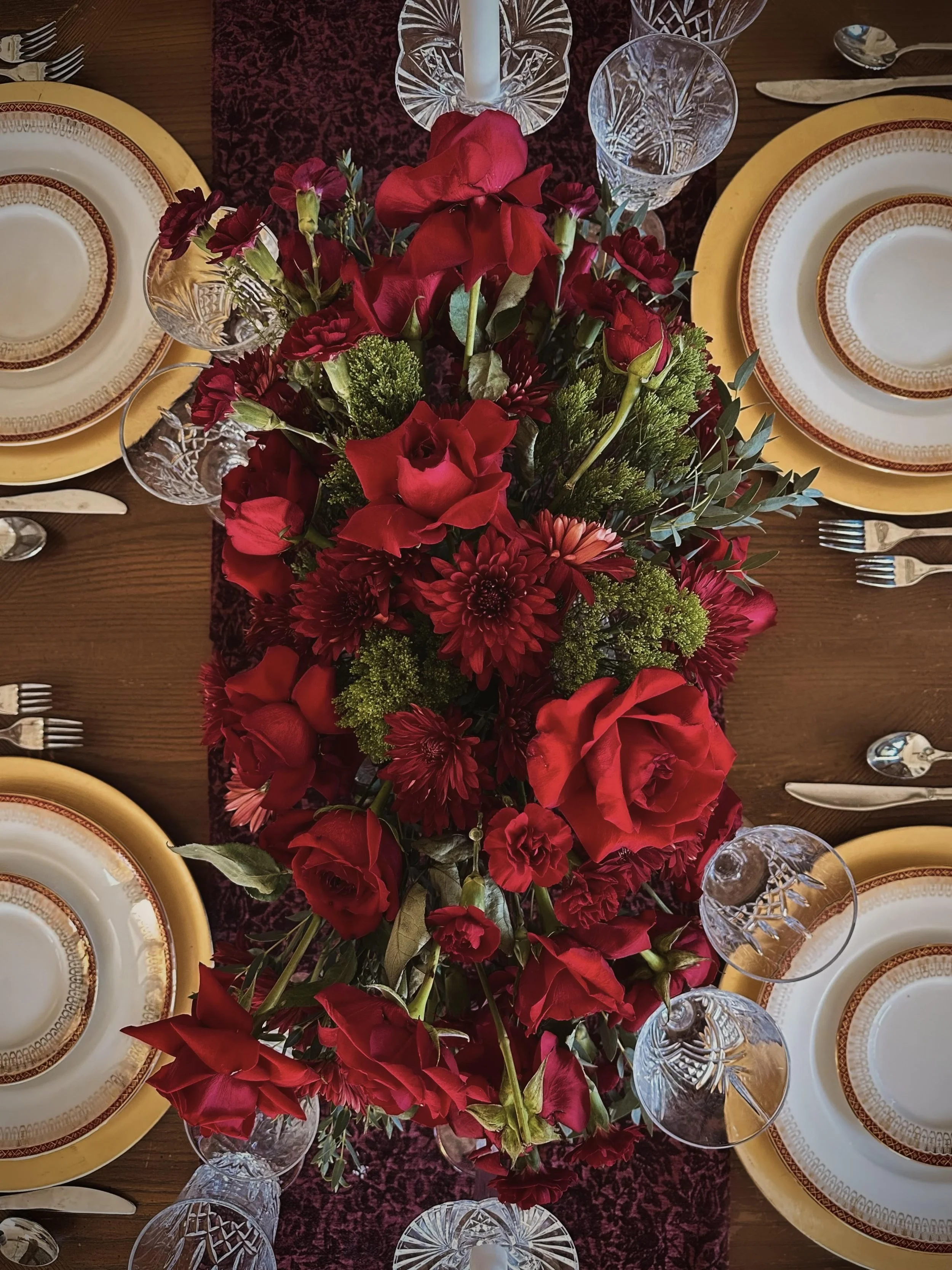 A dining table set with four place settings, featuring yellow plates with red and gold patterns, crystal glasses, silverware, and a burgundy table runner. In the center, there's an elaborate floral arrangement with red roses, dahlias, carnations, green foliage, and a large red bow.