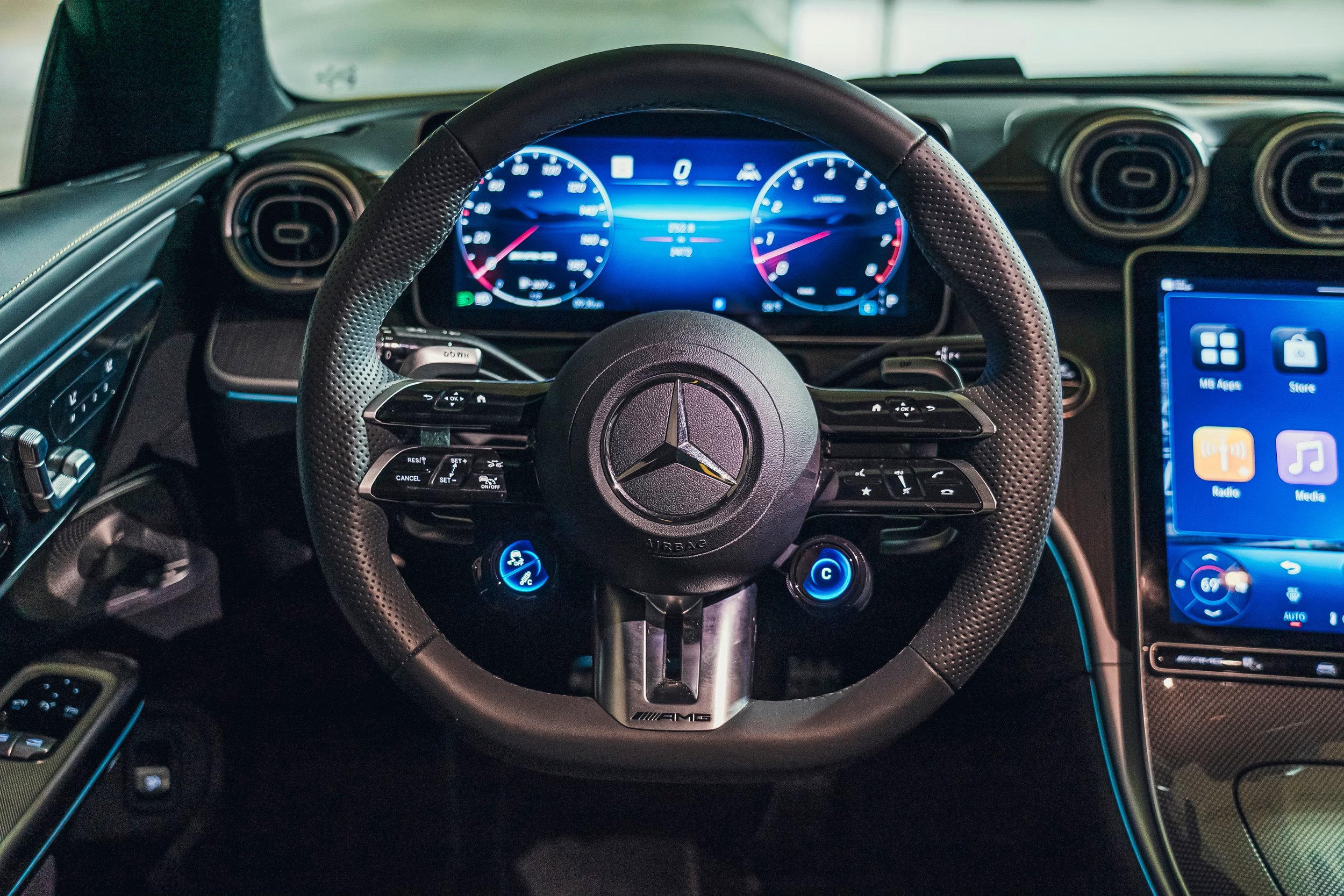 Interior of a Mercedes-Benz car, showing a leather-wrapped steering wheel with the Mercedes logo, a digital dashboard display, and an infotainment screen on the right side.