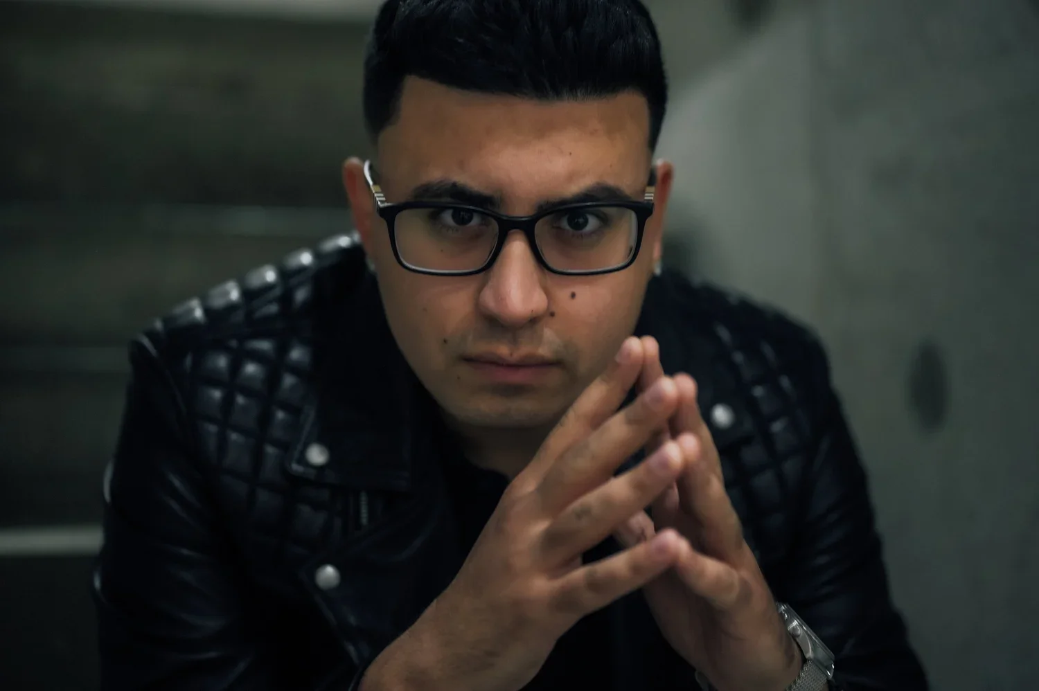 A young man with glasses, wearing a black leather jacket, looking directly at the camera with hands clasped together.