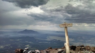 A landscape view from a mountain summit with a cloudy sky, distant mountains, and a weathered wooden signpost.