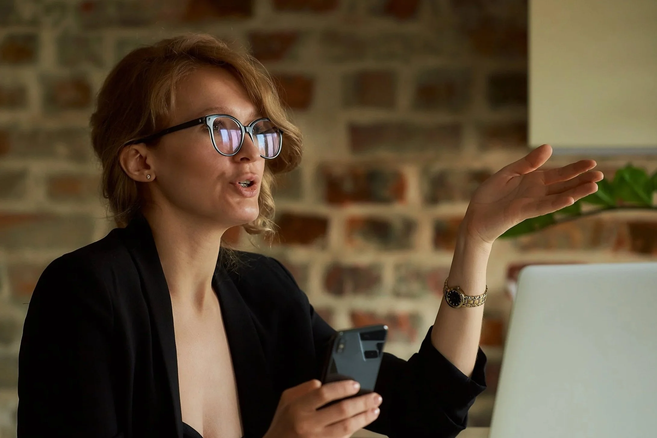 A woman with glasses and earrings in a black blazer is speaking while holding a smartphone in one hand and gesturing with her other hand near her head, seated in front of a laptop with a brick wall background.