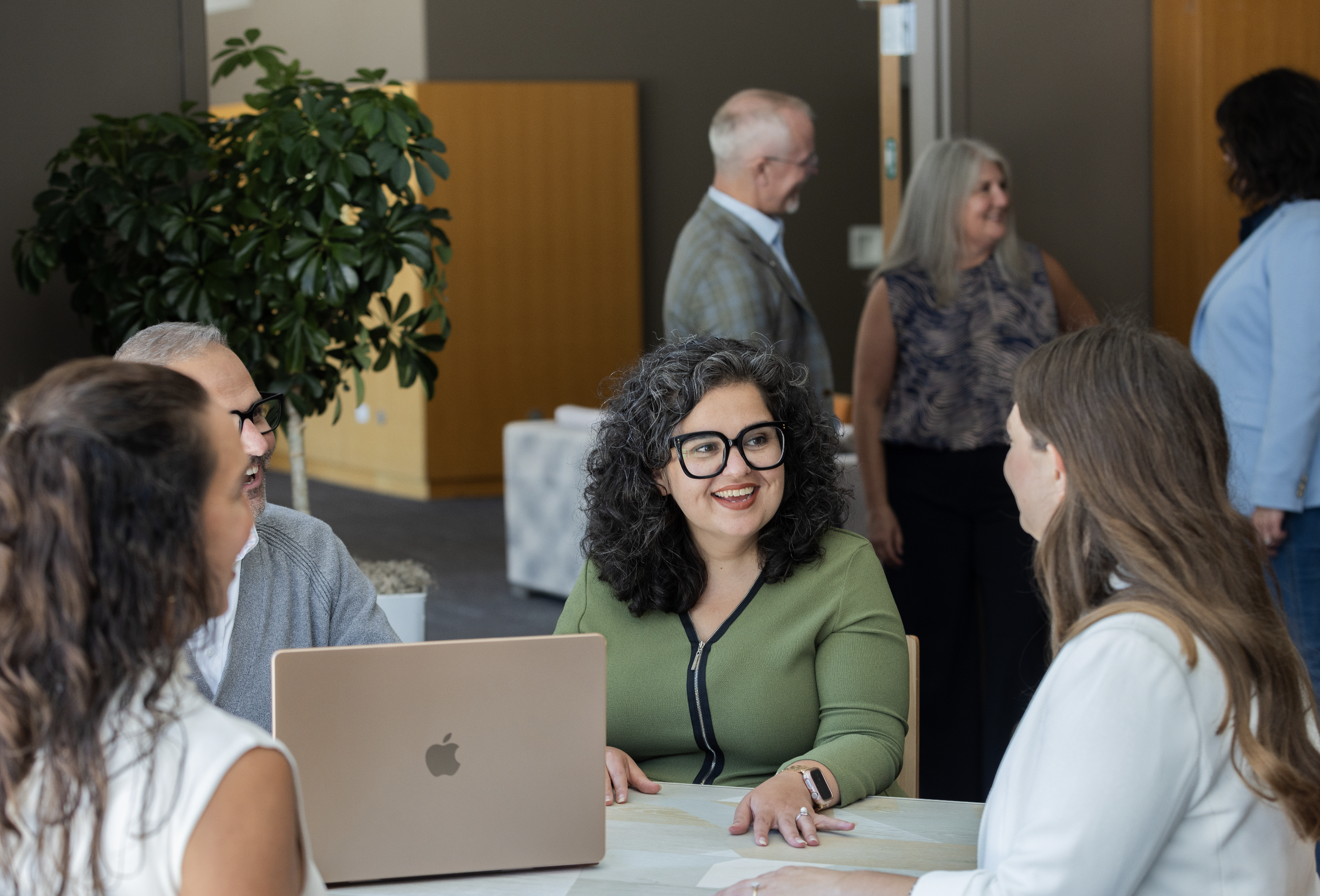 A group of five people in a professional setting engaging in a conversation indoors, with a woman with curly gray hair and glasses smiling at the center, and a laptop on the table.