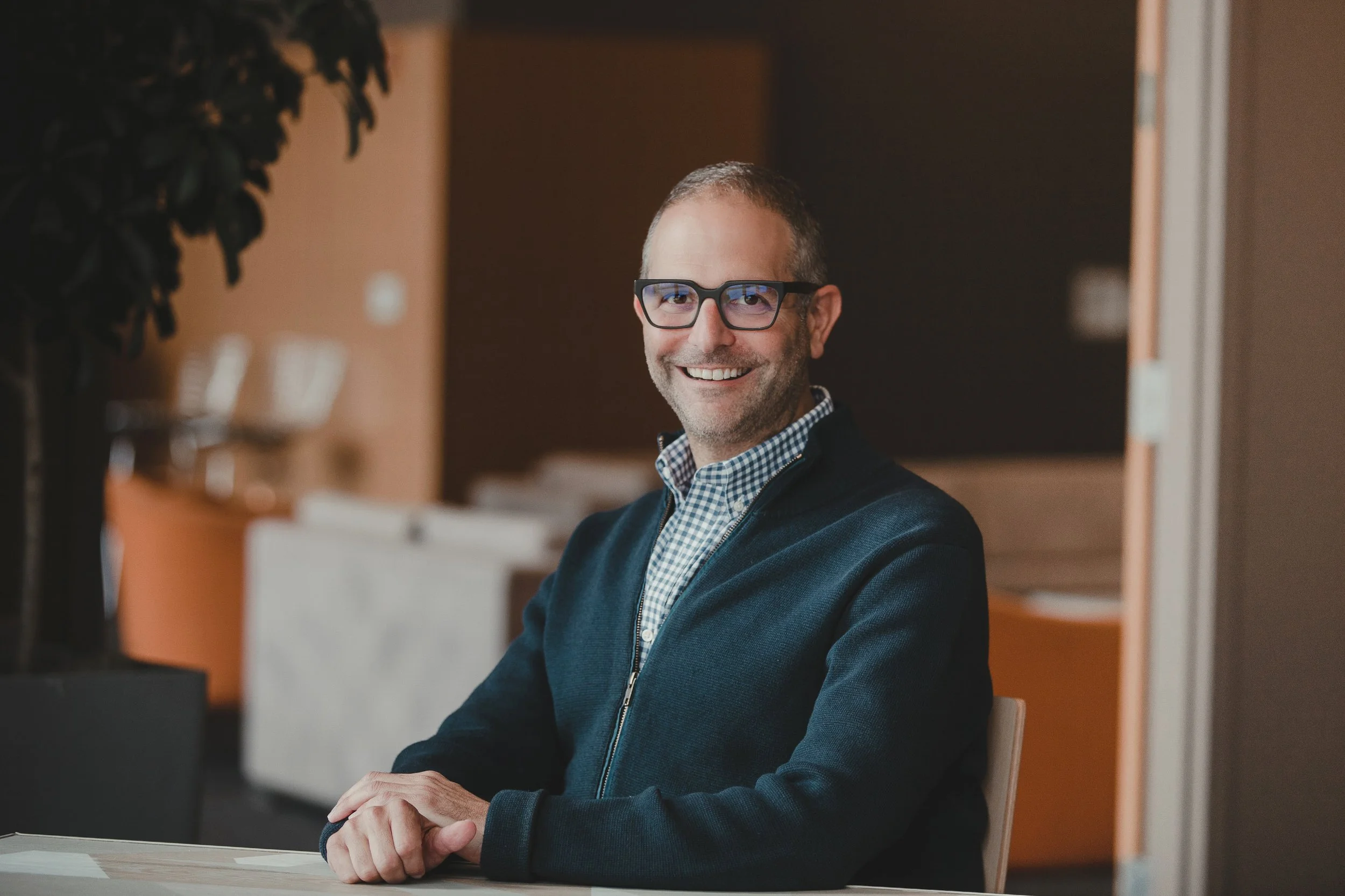 David, a smiling man with glasses and a beard sitting at a table in an office or waiting area.