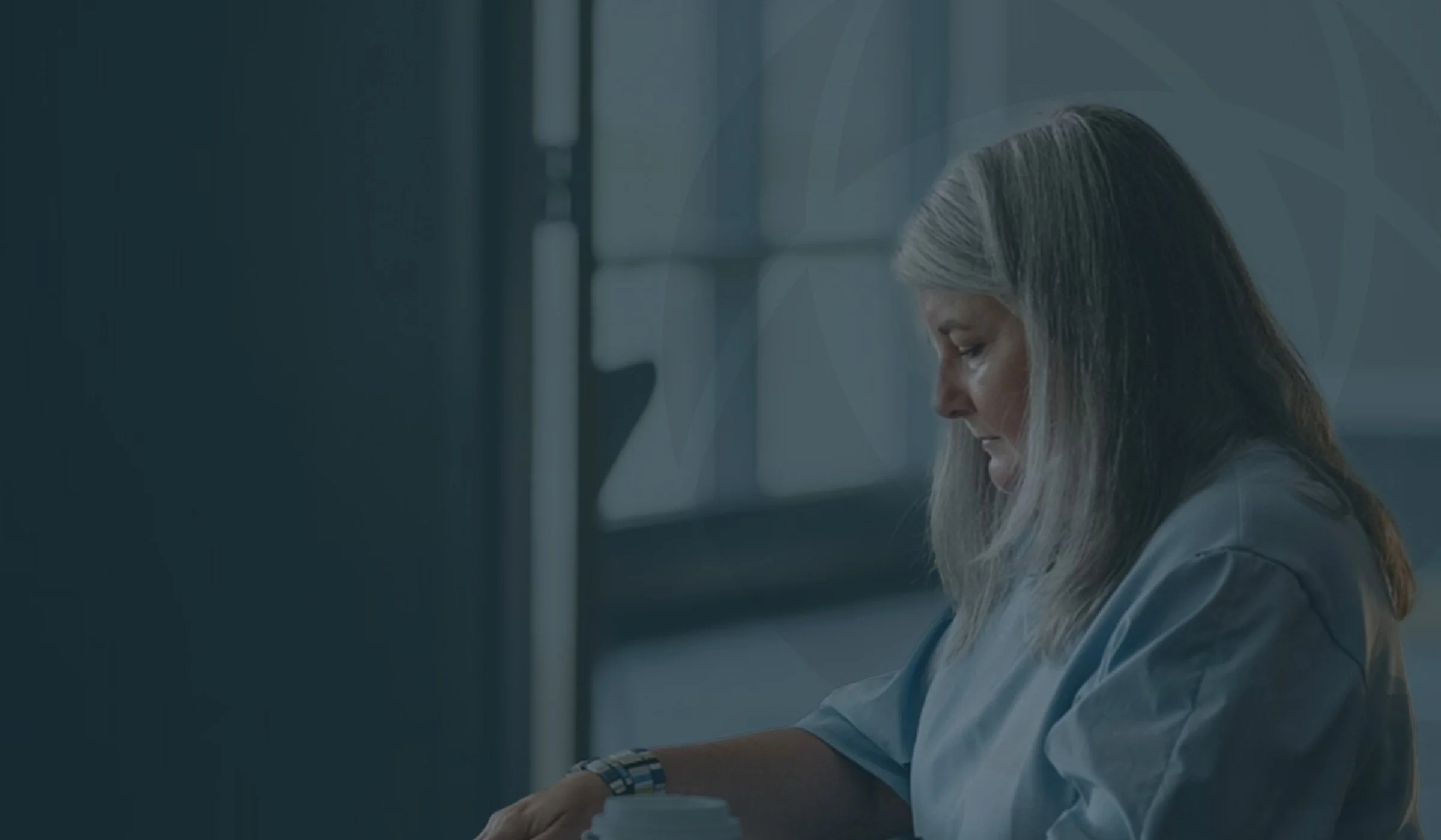 An elderly woman with gray hair sits at a table in a hospital gown, looking down pensively with a coffee cup in front of her, in a room with large windows.