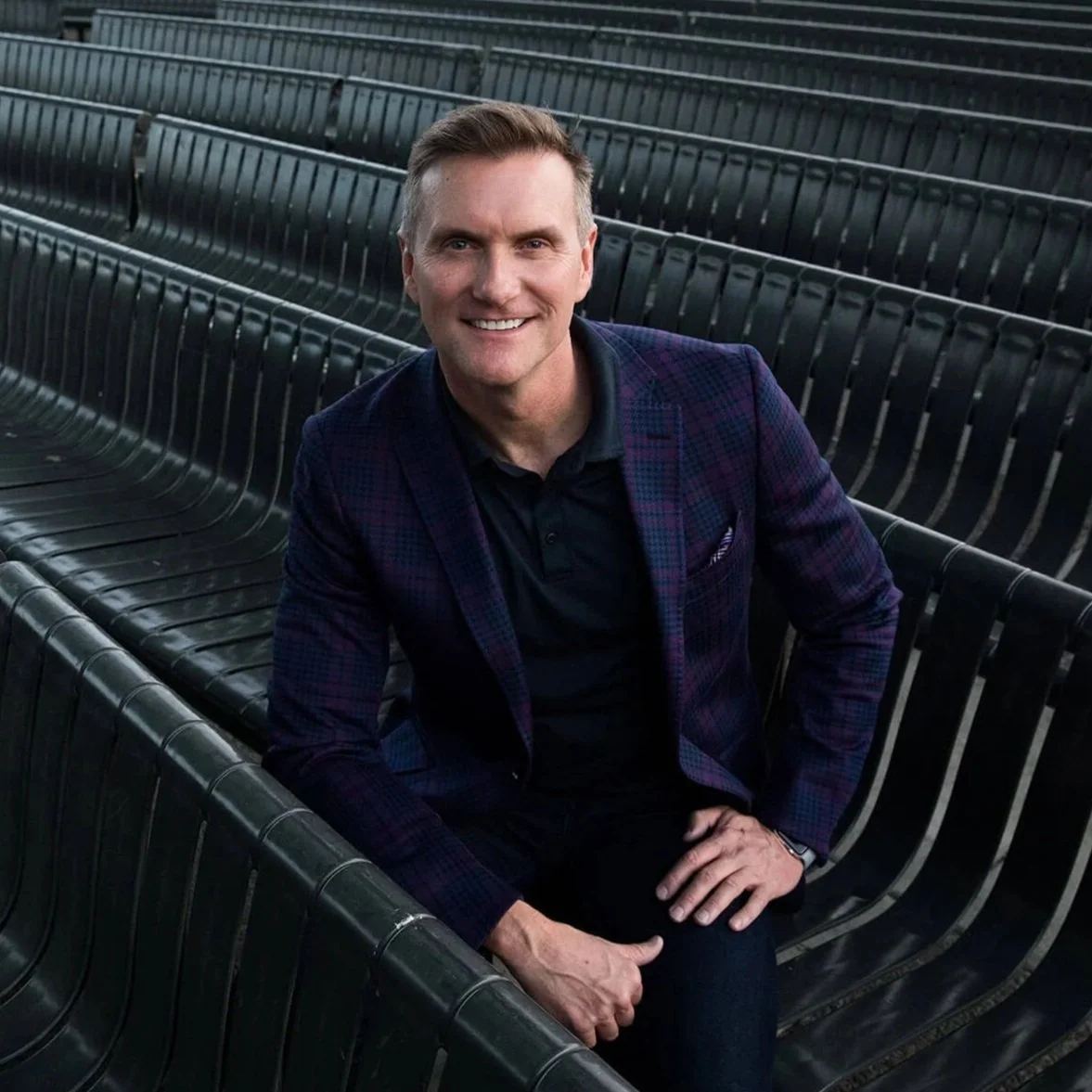 A smiling man, Buck Davis, with short hair, wearing a dark shirt and a purple checkered blazer, sitting on black stadium seats with a modern, minimalistic design.