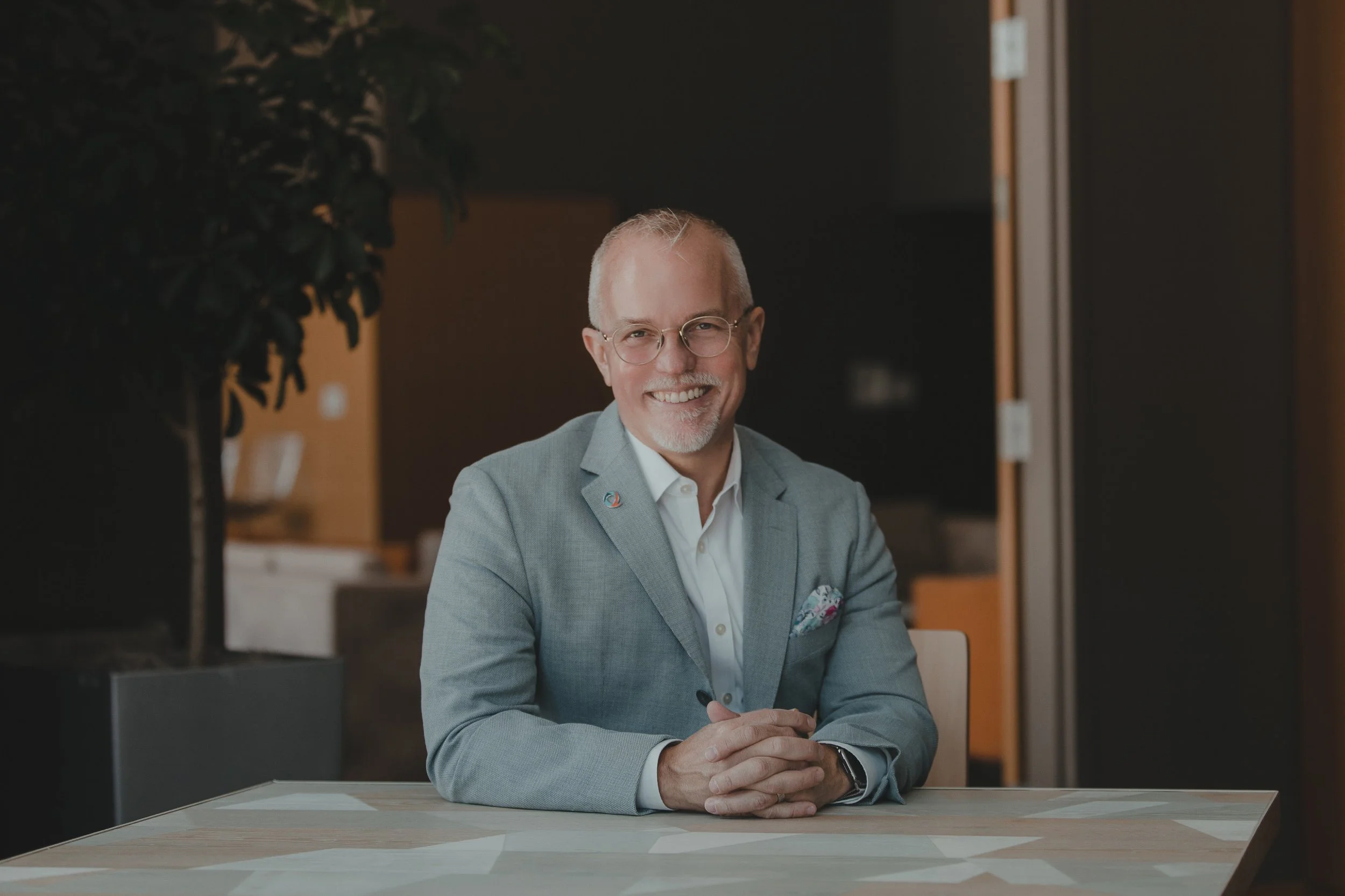 A smiling man, Scott Hoesman, in glasses and a light gray suit sitting at a table in a modern office environment.