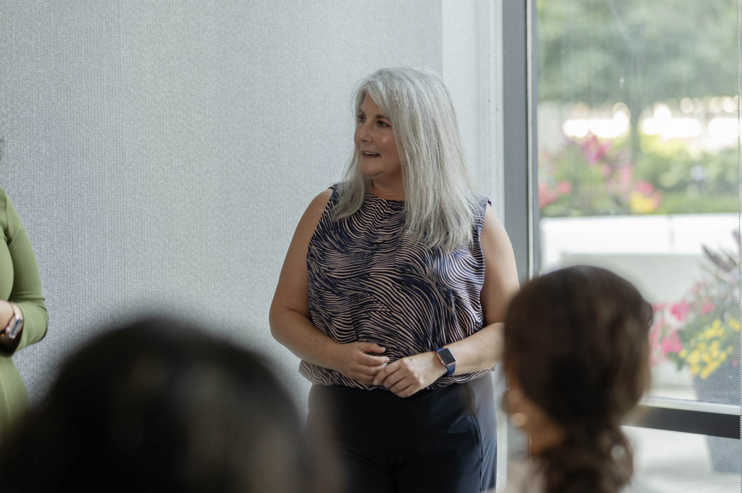Woman with gray hair wearing a sleeveless patterned top and a smartwatch, standing in front of a window and speaking to a group.