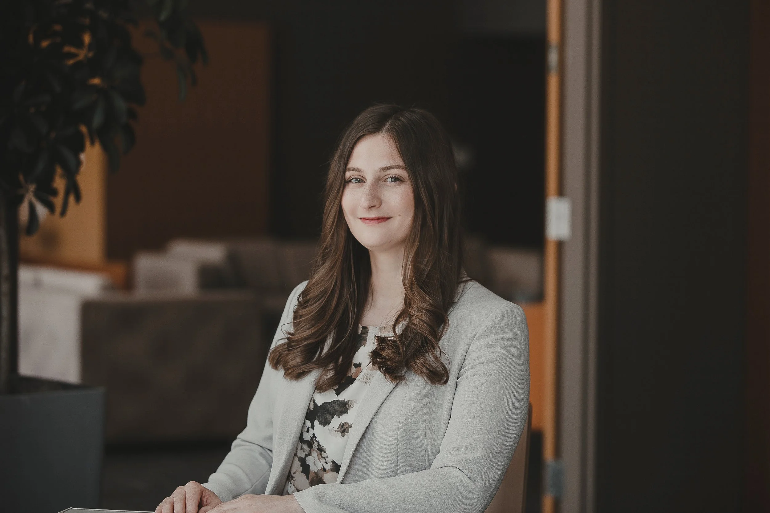 A woman wearing a light gray blazer and a floral top, sitting at a table in an indoor setting, smiling slightly and looking at the camera.