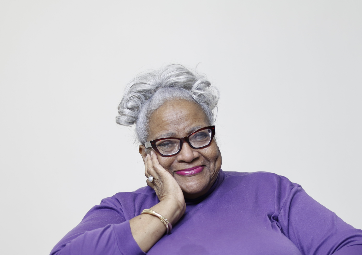 Gloria Cotton, an elderly woman with gray curly hair and glasses, smiling gently with her hand resting on her face, wearing a purple top.