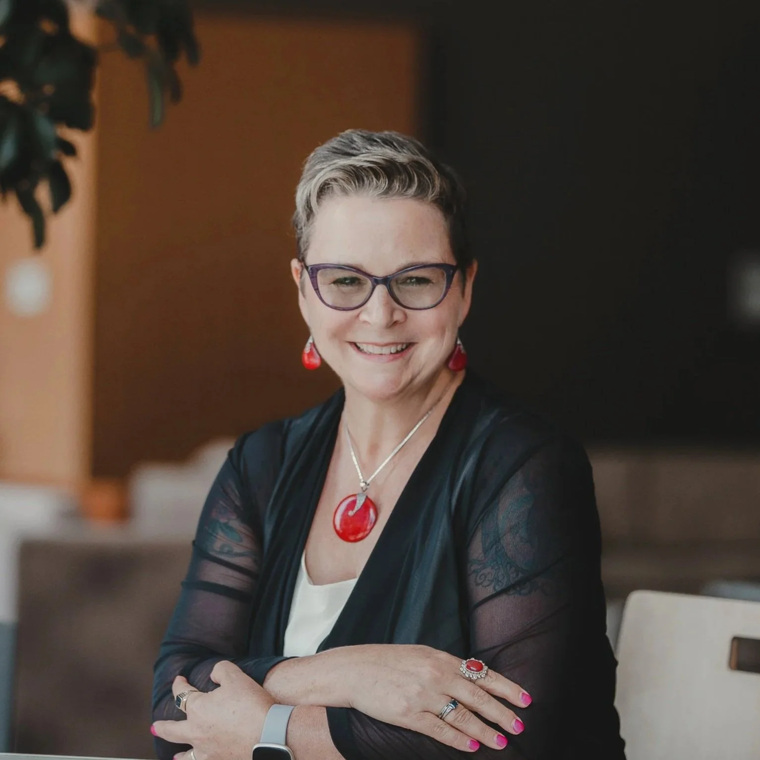 Kate Website, a smiling woman wearing glasses, red jewelry, and a black jacket with sheer sleeves, sitting at a table.