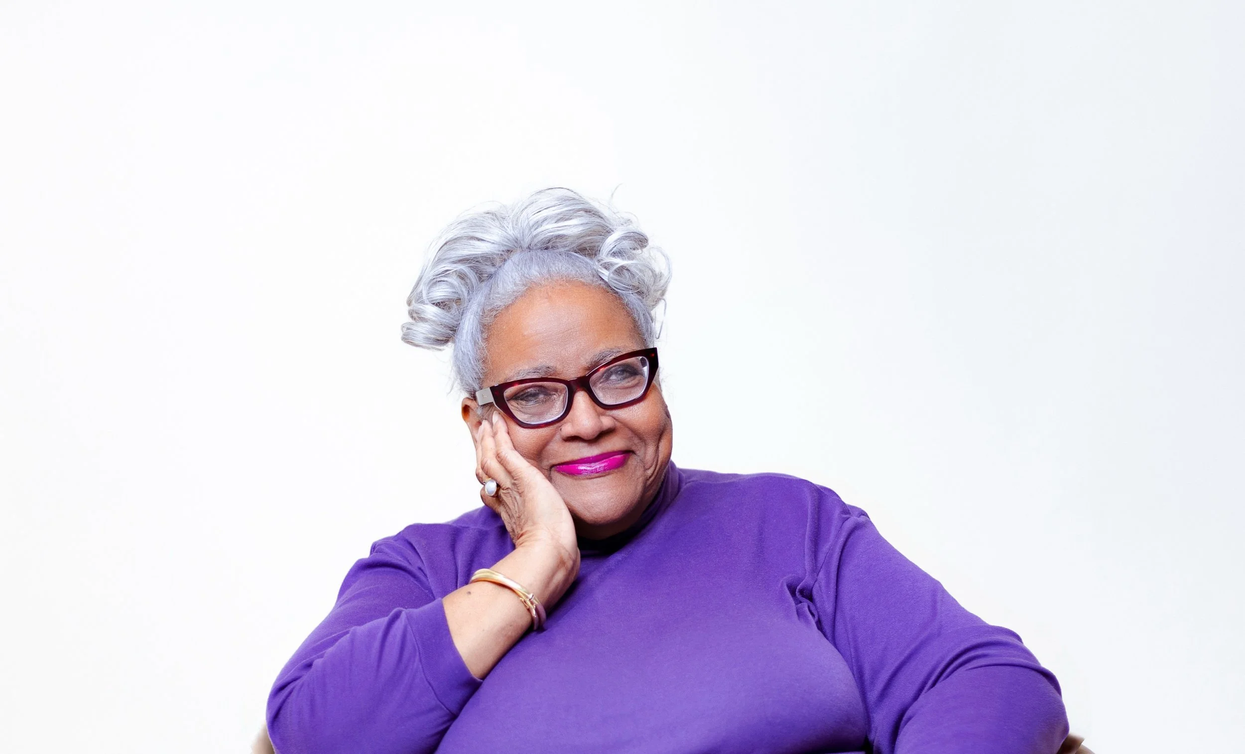 Gloria Cotton, a smiling elderly black woman with gray curly hair, wearing glasses, a purple top, and jewelry, resting her face on her hand against a plain white background.