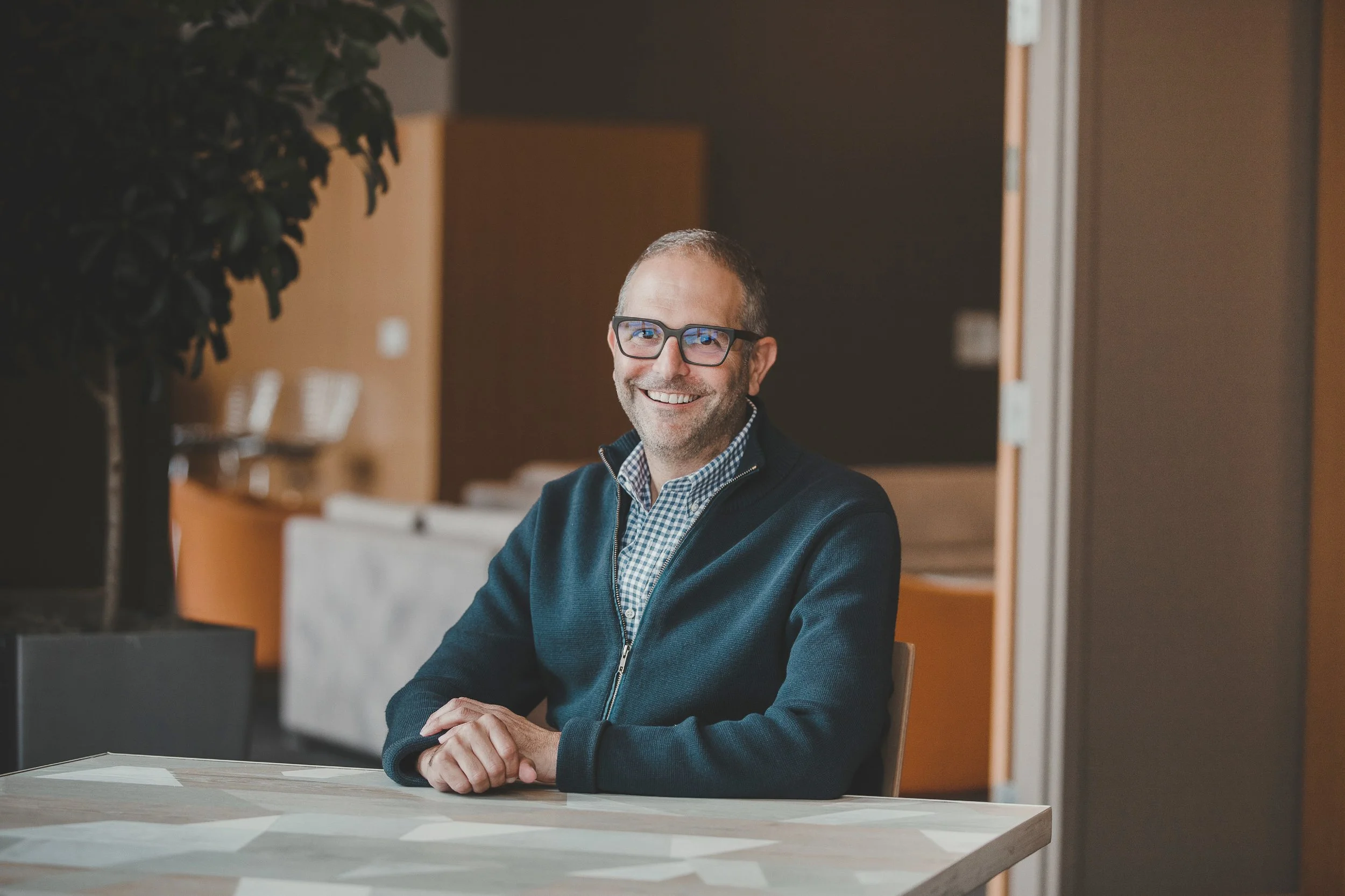 A man, David Stone,  with glasses, smiling, sitting at a table in a modern indoor setting, wearing a checkered shirt and dark jacket.