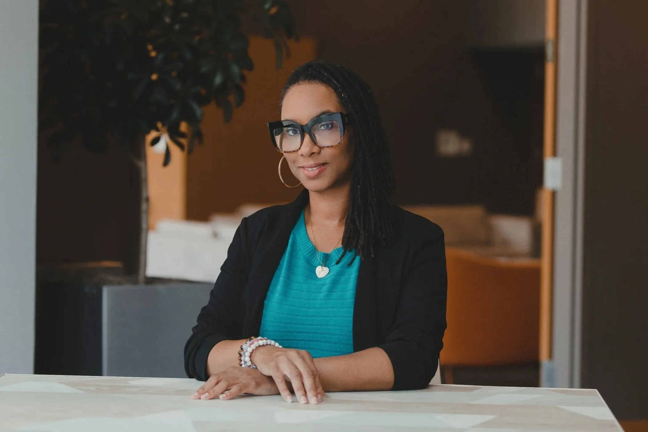 A woman, Danata Andrews, with black dreadlocks wearing large black glasses, hoop earrings, a turquoise top, a black blazer, and jewelry sitting at a table in a modern indoor setting.