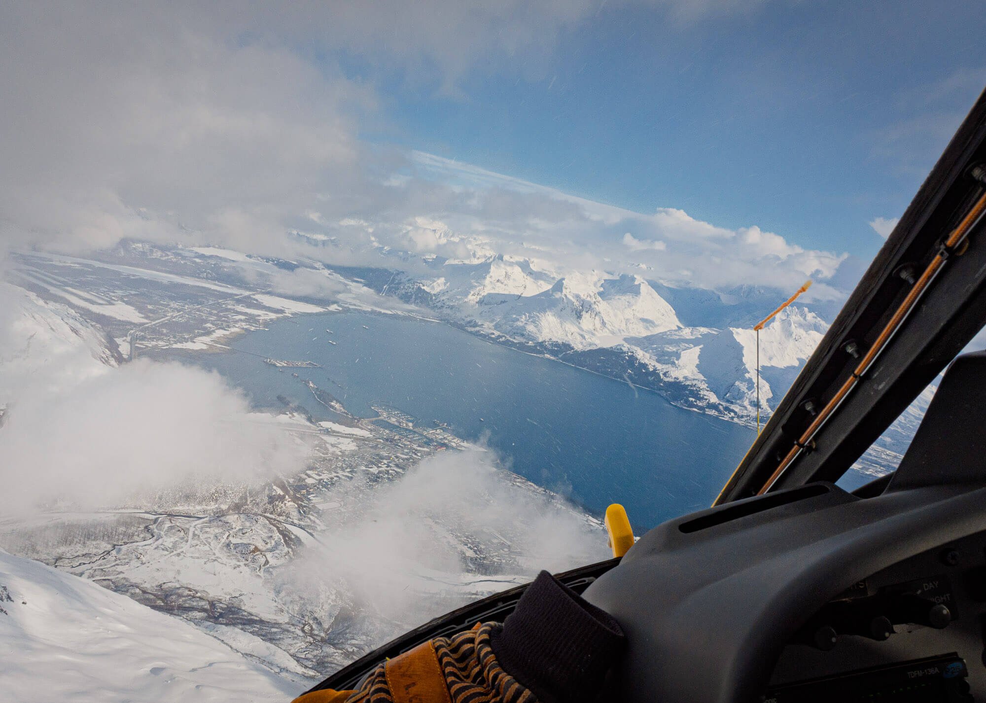 View from an airplane cockpit showing snow-covered mountains, a frozen lake, and cloudy skies.