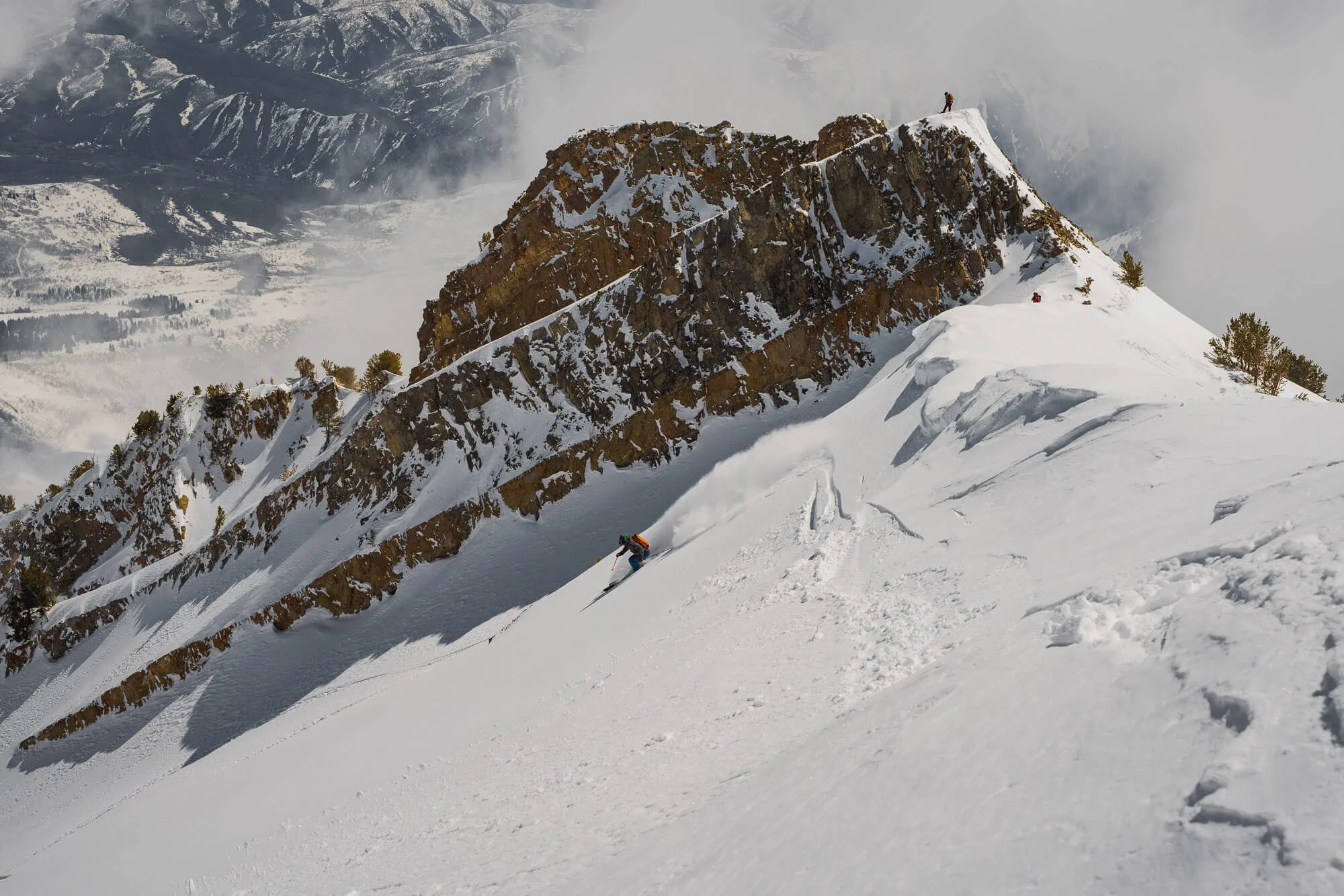 Mountaineer climbing snow-covered mountain with rocky cliffs, with distant snowy hills and clouds in the background.