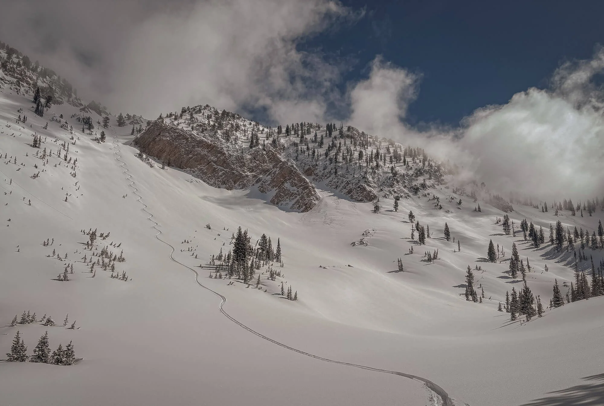 Snow-covered mountain slope with scattered pine trees, a winding ski trail, and rocky cliffs under a cloudy sky.
