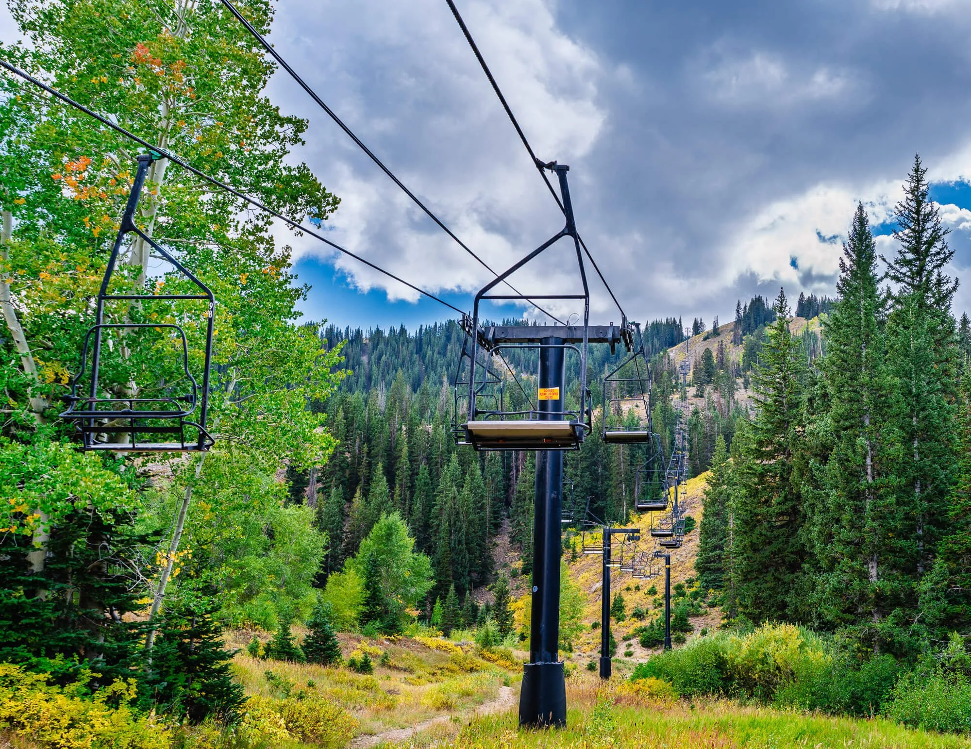 Empty ski lift chairs on a mountain surrounded by green trees and cloudy sky.