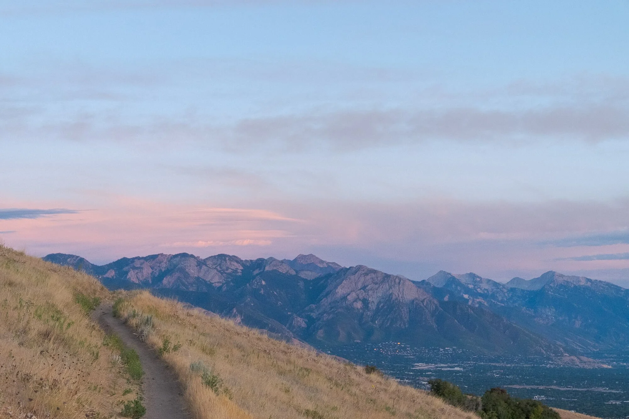 A mountain landscape at dusk with a dirt trail on a grassy hillside leading towards a range of tall mountains under a pink and blue sky.