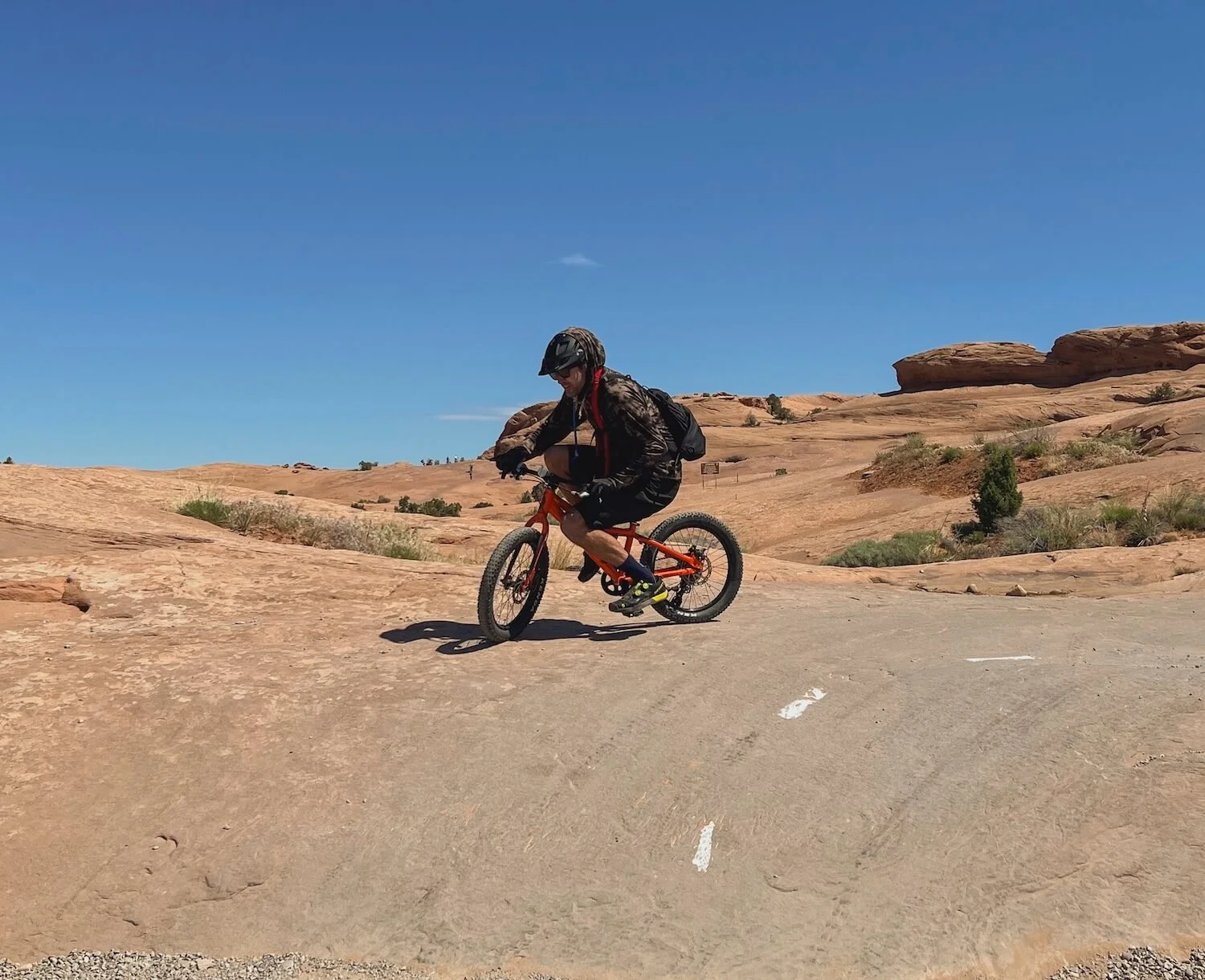A person wearing a helmet and dark clothing riding a mountain bike on a rocky trail in a desert landscape with sparse vegetation and red rock formations under a clear blue sky.