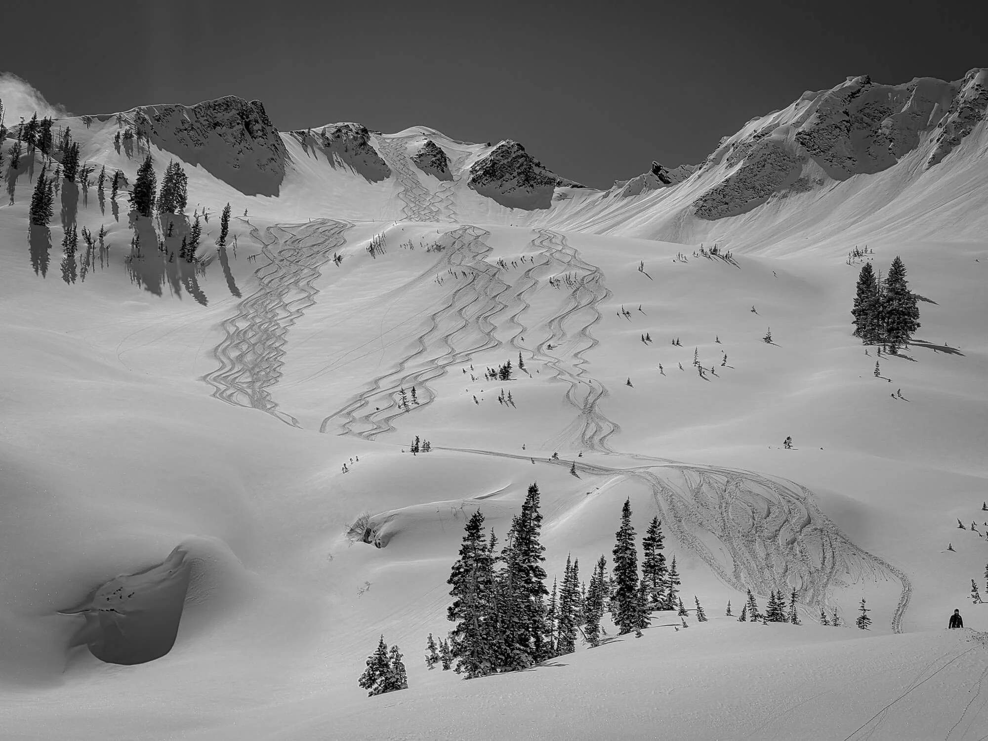 Snow-covered mountain landscape with ski tracks, pine trees, and rugged peaks under a partly cloudy sky.