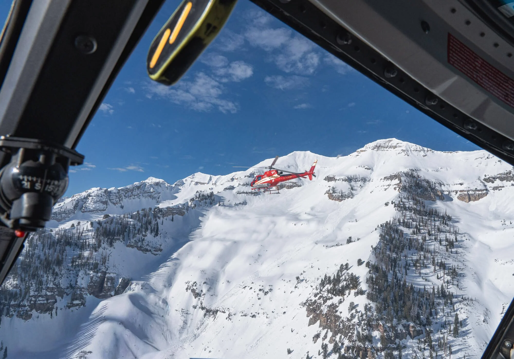 A red helicopter flying over snow-covered mountains, captured from inside another helicopter.