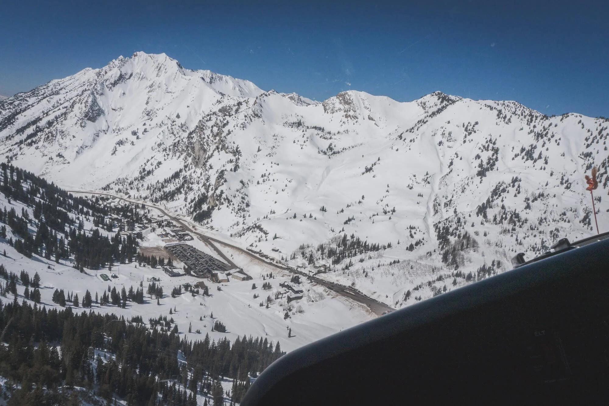 Snow-covered mountain landscape with a small town, forests, and winding roads, seen from a airplane cockpit.