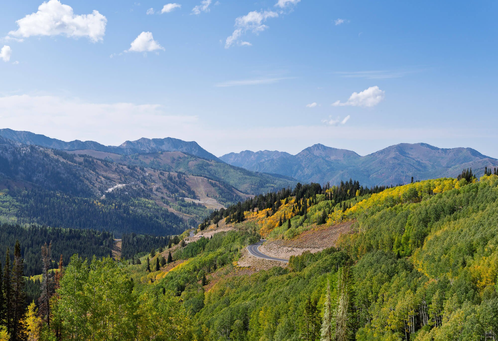 Scenic view of a winding road through lush green mountains with patches of yellow and orange trees, under a blue sky with scattered clouds.